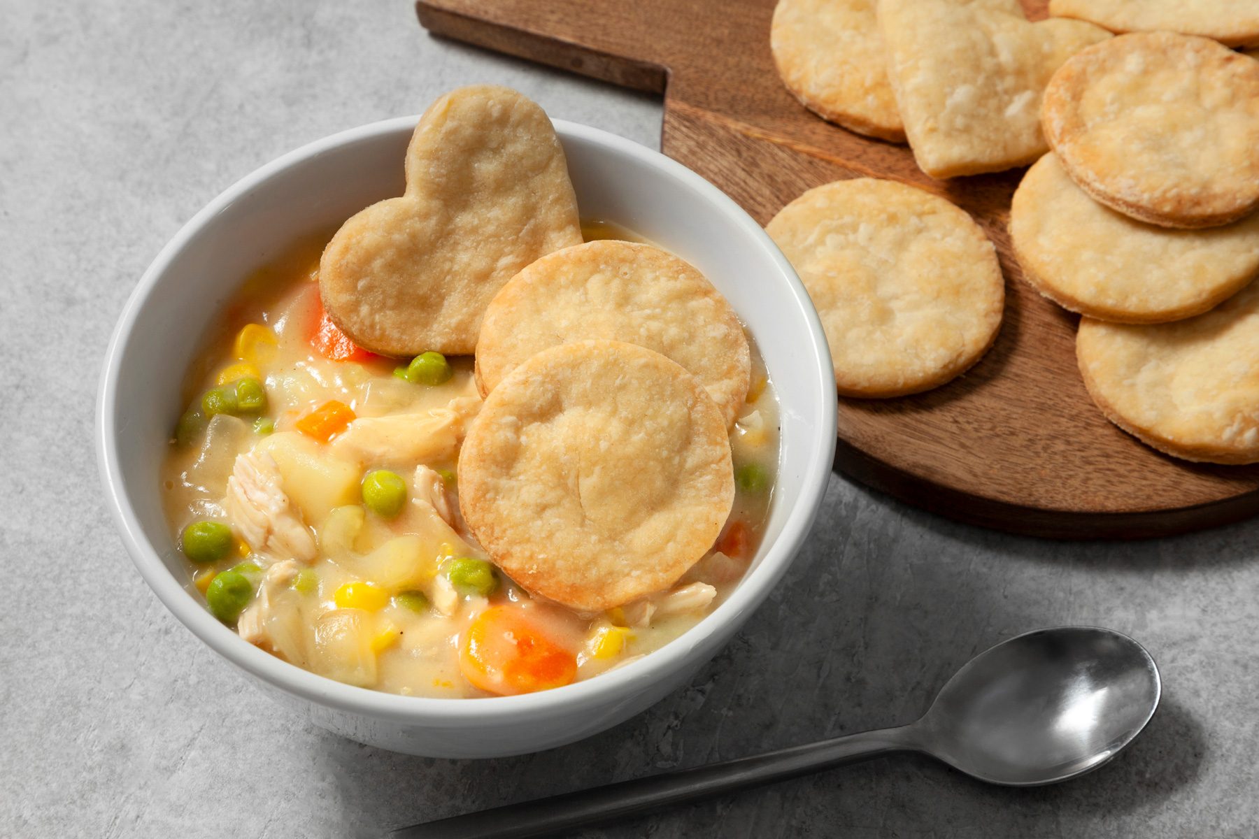 3/4th shot; light grey background; A bowl of Chicken Pot Pie Soup is featured, filled with tender chicken, vegetables, and a creamy broth; Heart-shaped biscuits are arranged on a wooden cutting board and also added over the dish