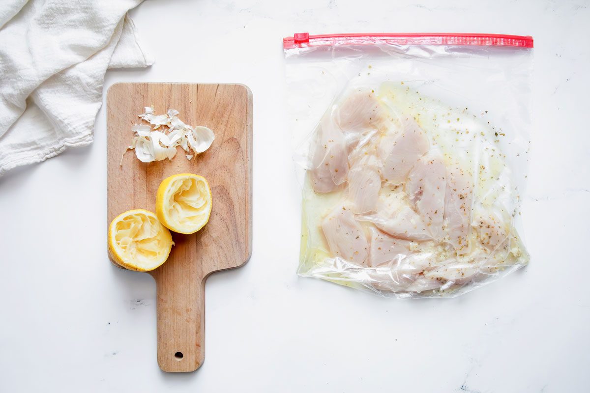 Overhead shot for Taste of Home Chicken Gyros with chicken marinating in a large ziplock bag and used ingredients on a wooden cutting board on a marble surface.
