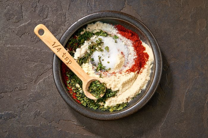 A black ceramic bowl filled with a mixture of dried herbs and spices, featuring green parsley, red paprika, white salt, and cream-colored flour. A wooden measuring spoon labeled "1/4 tsp" rests on top, partially buried in the ingredients. Dark textured background.