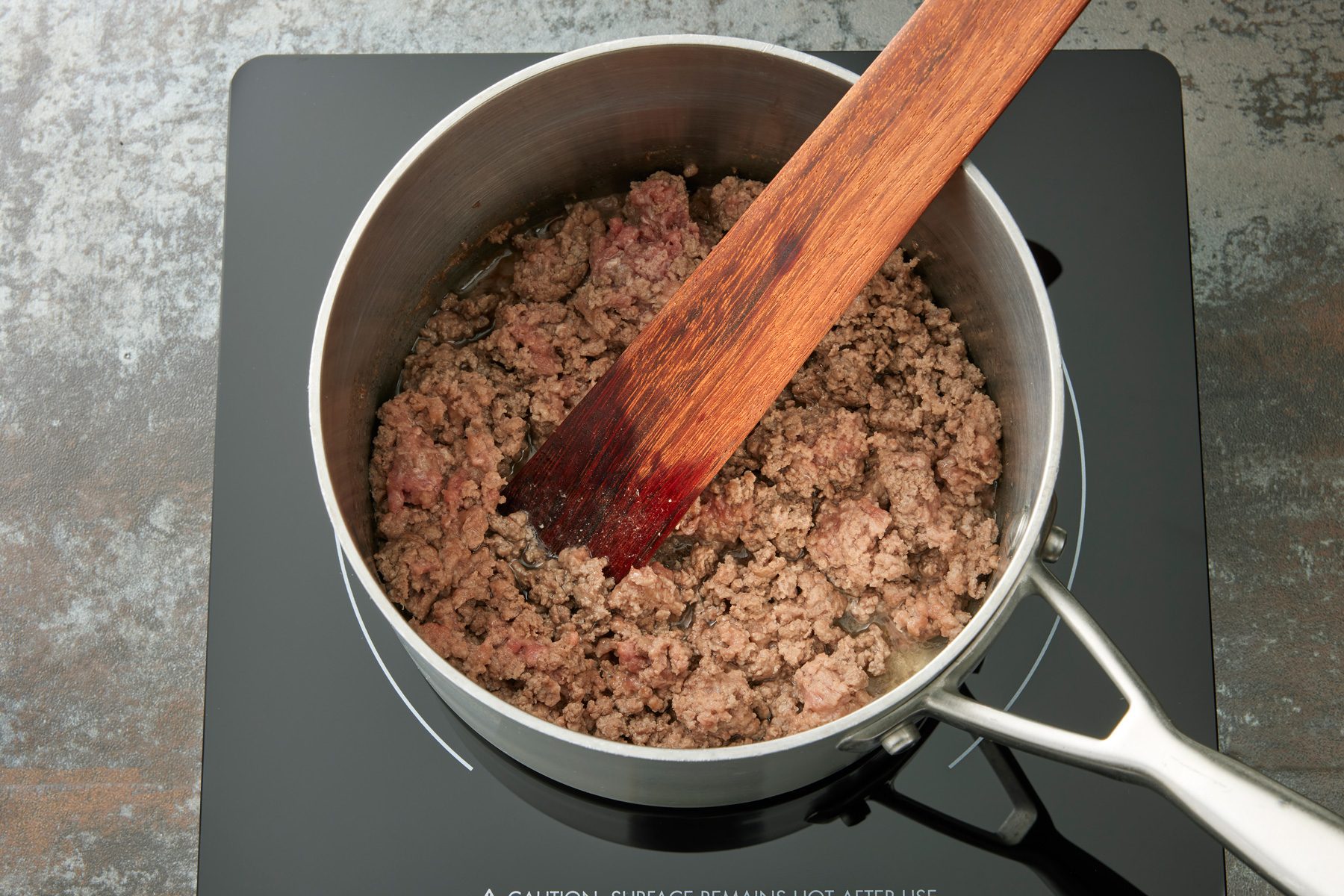 Overhead shot of in a large saucepan over medium heat; cook beef until no longer pink; breaking into crumbles; drain; wooden flat spoon; induction; dark texture surface