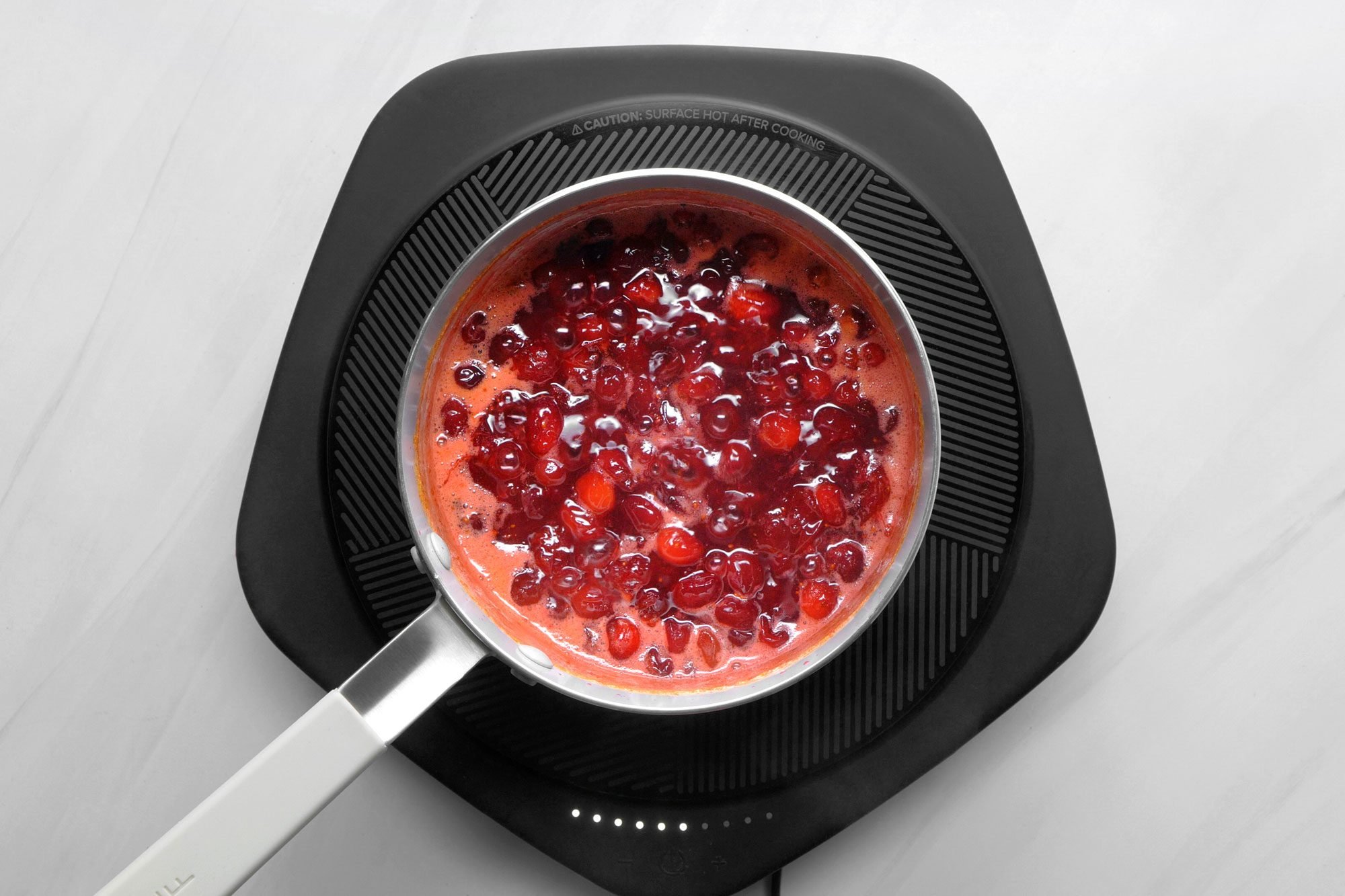 overhead shot; white background; A saucepan on an electric induction cooktop contains a mixture of cranberries and a liquid, likely orange juice or sugar syrup, The cranberries are a vibrant red color and are simmering in the liquid; The sauce is thick and appears to be bubbling gently;