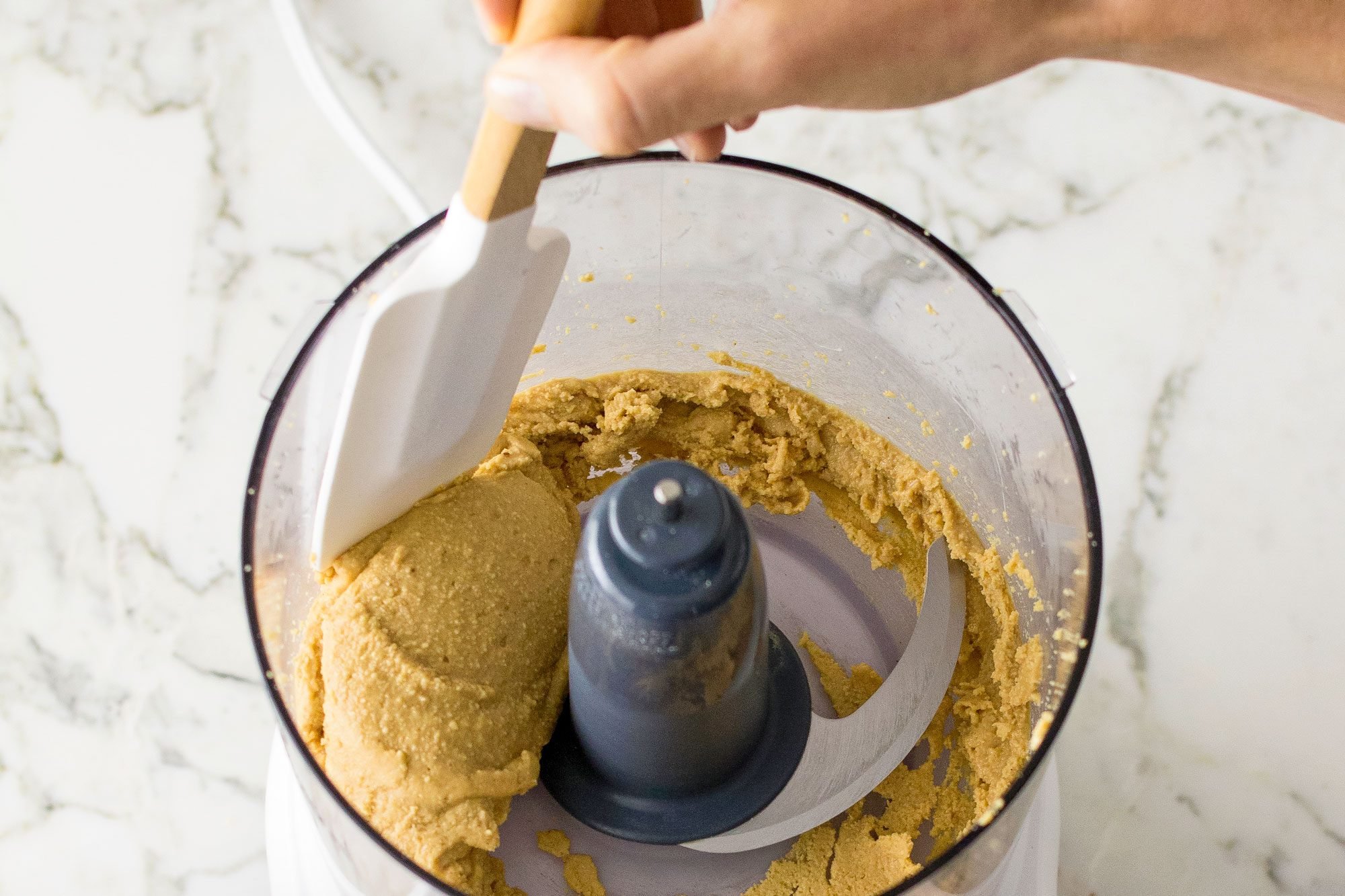Blending Peanuts in a food processor; marble background; spatula in hand;