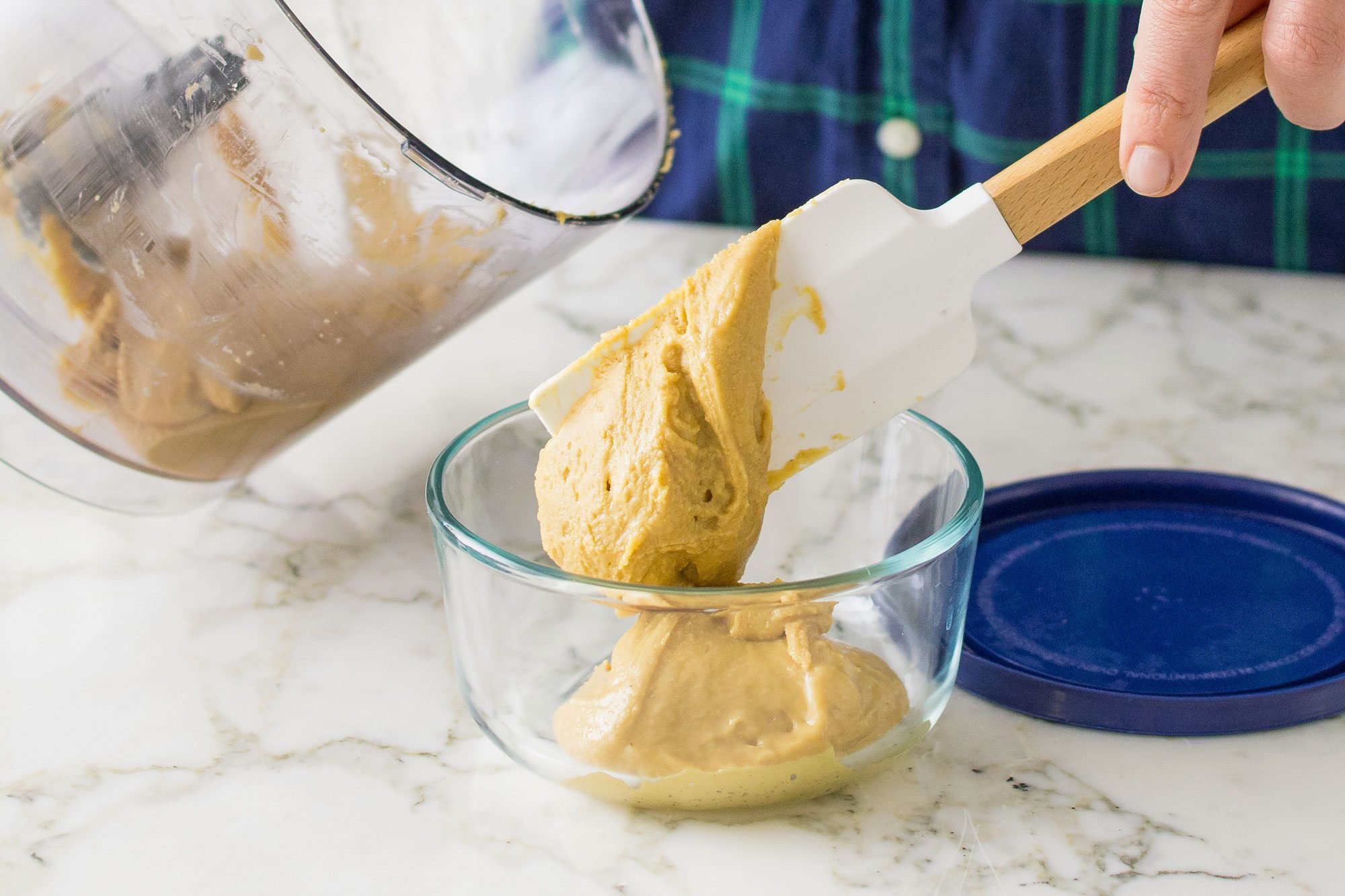 transferring peanut butter from food processor to glass container with spatula; marble background;
