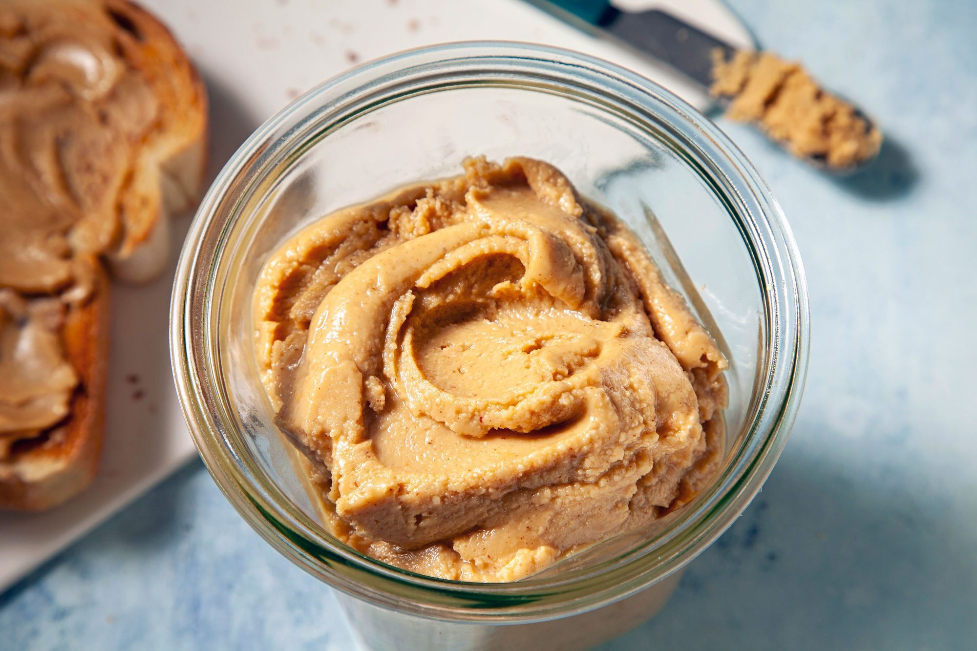 3/4th shot; light blue textured background; A glass jar filled with creamy peanut butter takes center stage; A slice of bread with peanut butter spread on it is visible in the background;