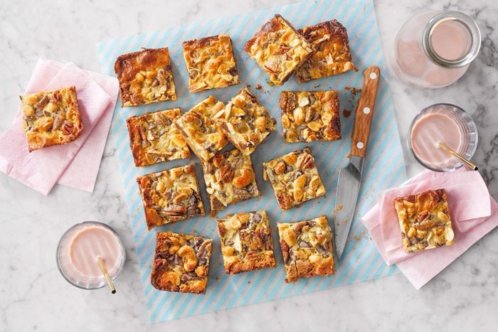 A marble countertop features a blue-striped baking sheet with several pieces of nut and dried fruit-topped bar cookies. Two glasses of chocolate milk with straws and a knife are nearby, along with napkins holding additional cookie pieces.
