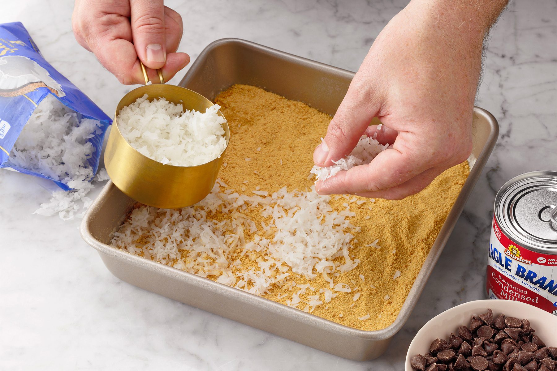 A person is sprinkling shredded coconut from a measuring cup onto a layer of crumbled graham crackers in a square baking pan. Surrounding the pan are baking ingredients including chocolate chips, shredded coconut, and a can of sweetened condensed milk.