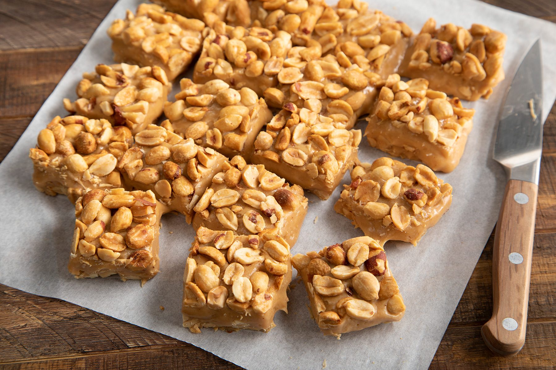 Squares of homemade peanut butter fudge, topped with whole peanuts, are neatly arranged on a sheet of parchment paper. A knife with a wooden handle rests beside them on a wooden surface.