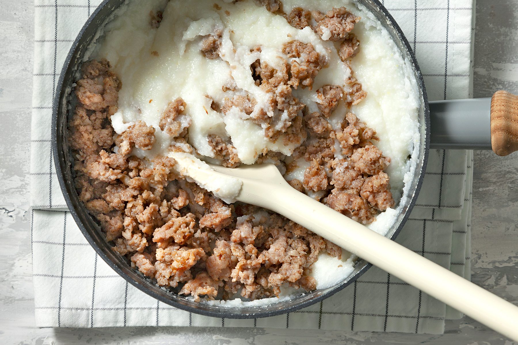 A frying pan filled with ground sausage and a white, creamy substance, possibly gravy. A white spatula is placed in the mixture, and the pan rests on a checkered kitchen towel on a light-textured countertop.