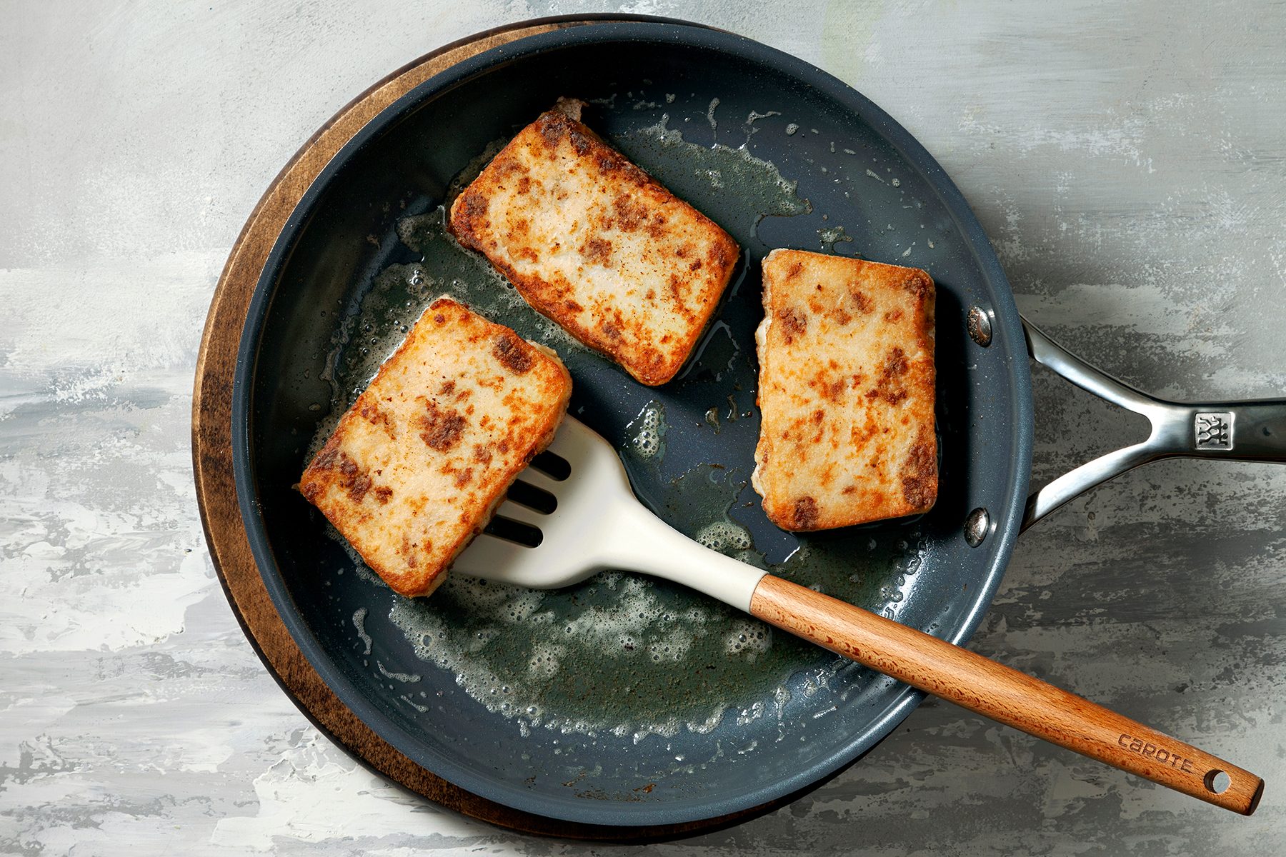 A frying pan with three rectangular pieces of food, likely fried tofu or a similar item, being cooked. A spatula with a wooden handle rests in the pan, lifting one piece. The pan sits on a textured gray surface.