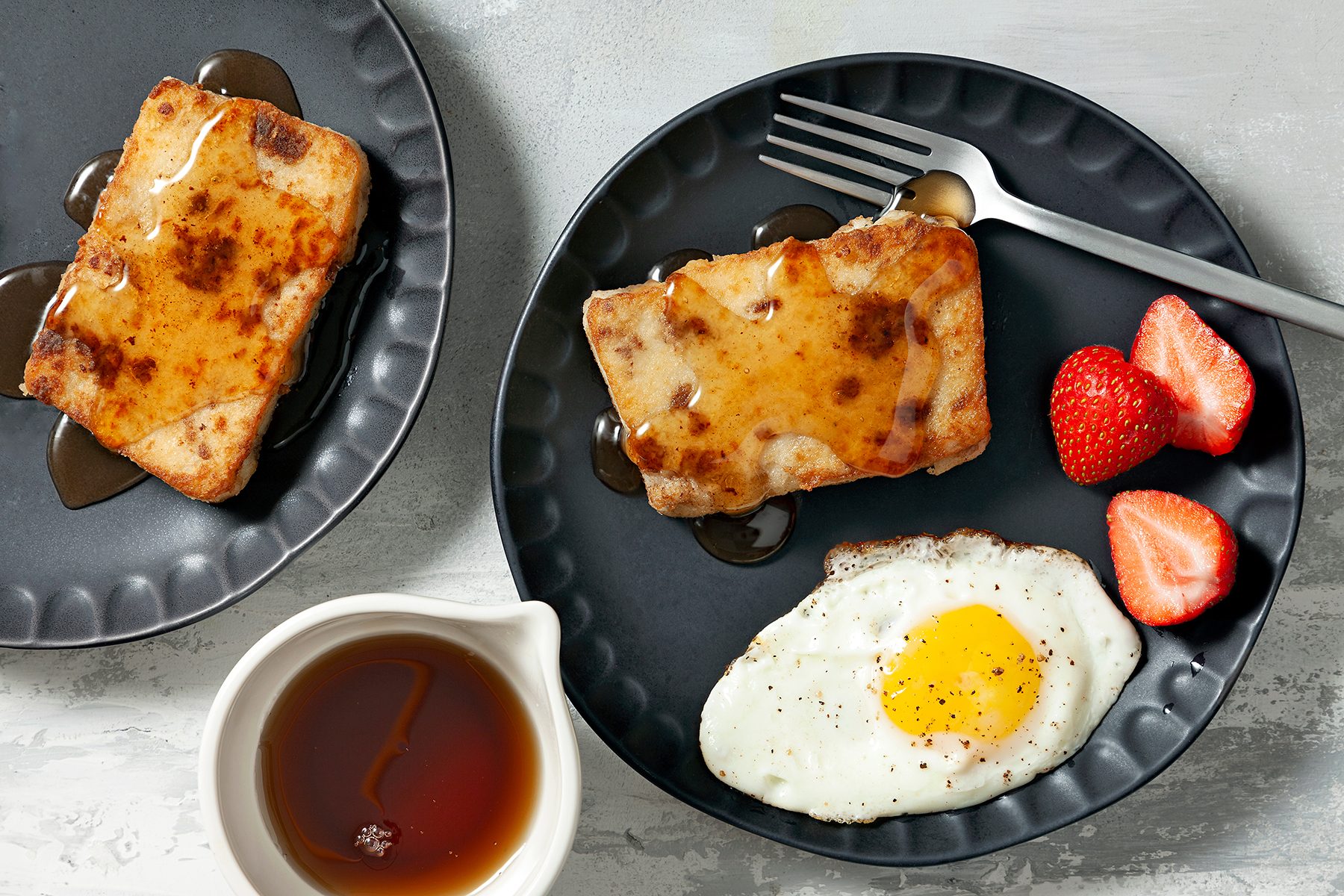 Two black plates each hold French toast topped with syrup. One plate includes a sunny-side-up egg and halved strawberries. A small white pitcher filled with syrup sits nearby. A fork rests on one of the plates.