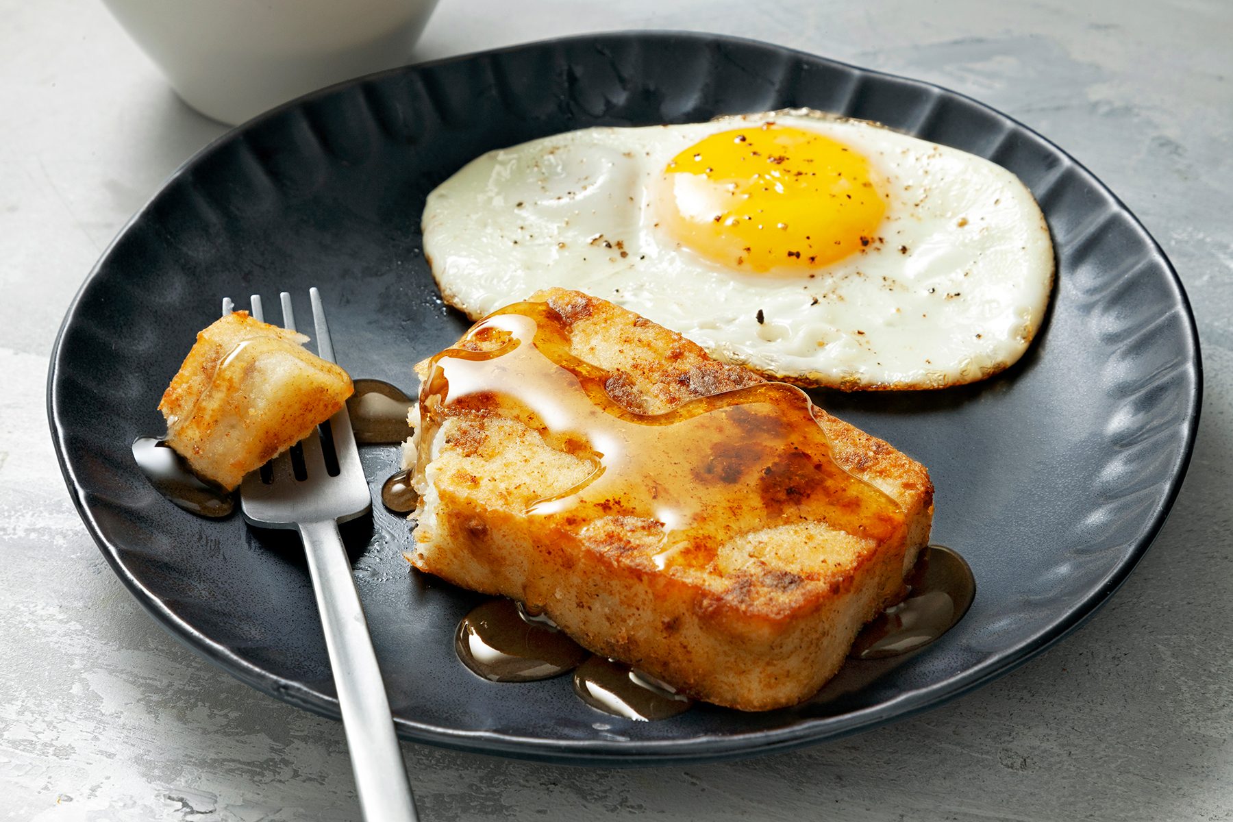 A black plate with a sunny-side-up egg seasoned with pepper and a thick slice of French toast topped with melted butter and syrup. A fork is placed on the plate with a piece of the toast speared on its tines.
