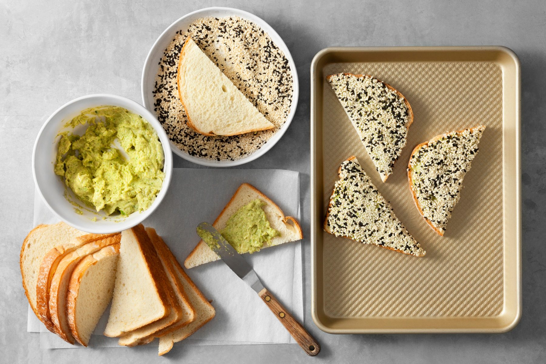 overhead shot; light grey background; A bowl of creamy shrimp mixture; Slices of white bread are ready to be topped with the mixture and coated in sesame seeds; A baking sheet lined with parchment paper awaits the assembled toasts