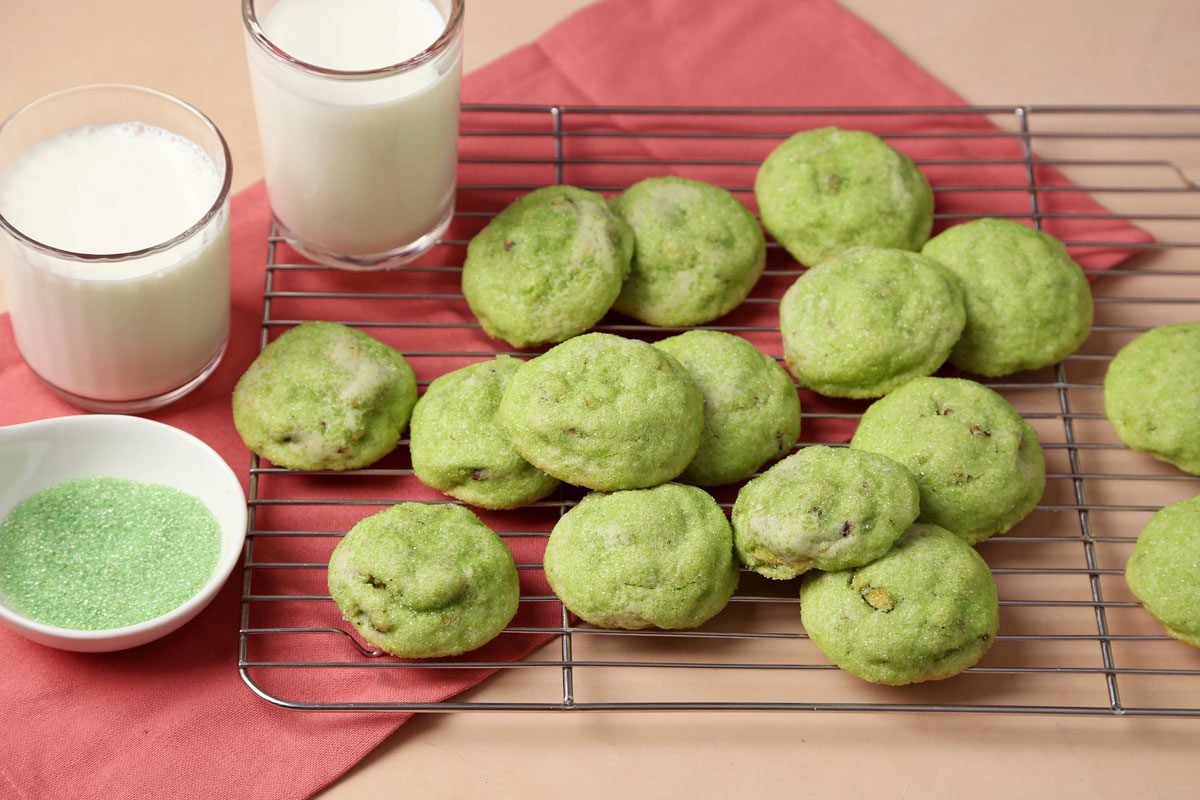 Close up of Taste of Home's Pistachio Cookies on a wire rack on a pink surface with glasses of milk.