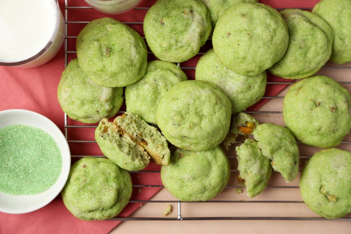 Close up of Taste of Home's Pistachio Cookies on a wire rack on a pink surface with glasses of milk.
