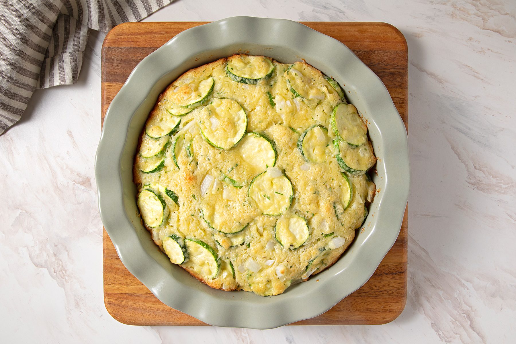 A zucchini casserole sits in a round, fluted dish atop a wooden board on a marble countertop. The surface of the casserole is golden and speckled with visible zucchini slices. A striped cloth is partially visible in the top left corner.