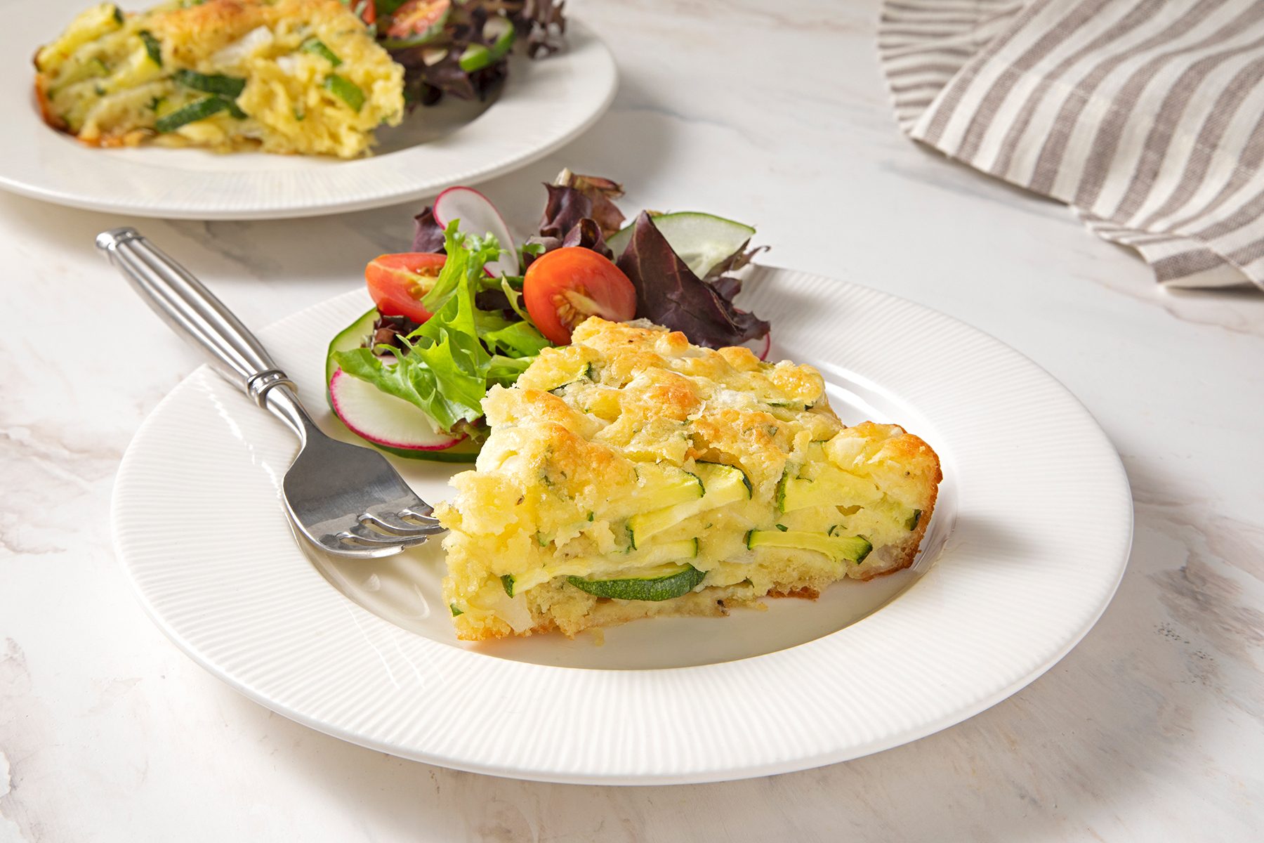 A slice of savory pie with zucchini and cheese on a white plate, accompanied by a fresh mixed greens salad with tomatoes and radishes. A fork rests on the plate. Another plate with the same dish and a striped cloth napkin are in the background.