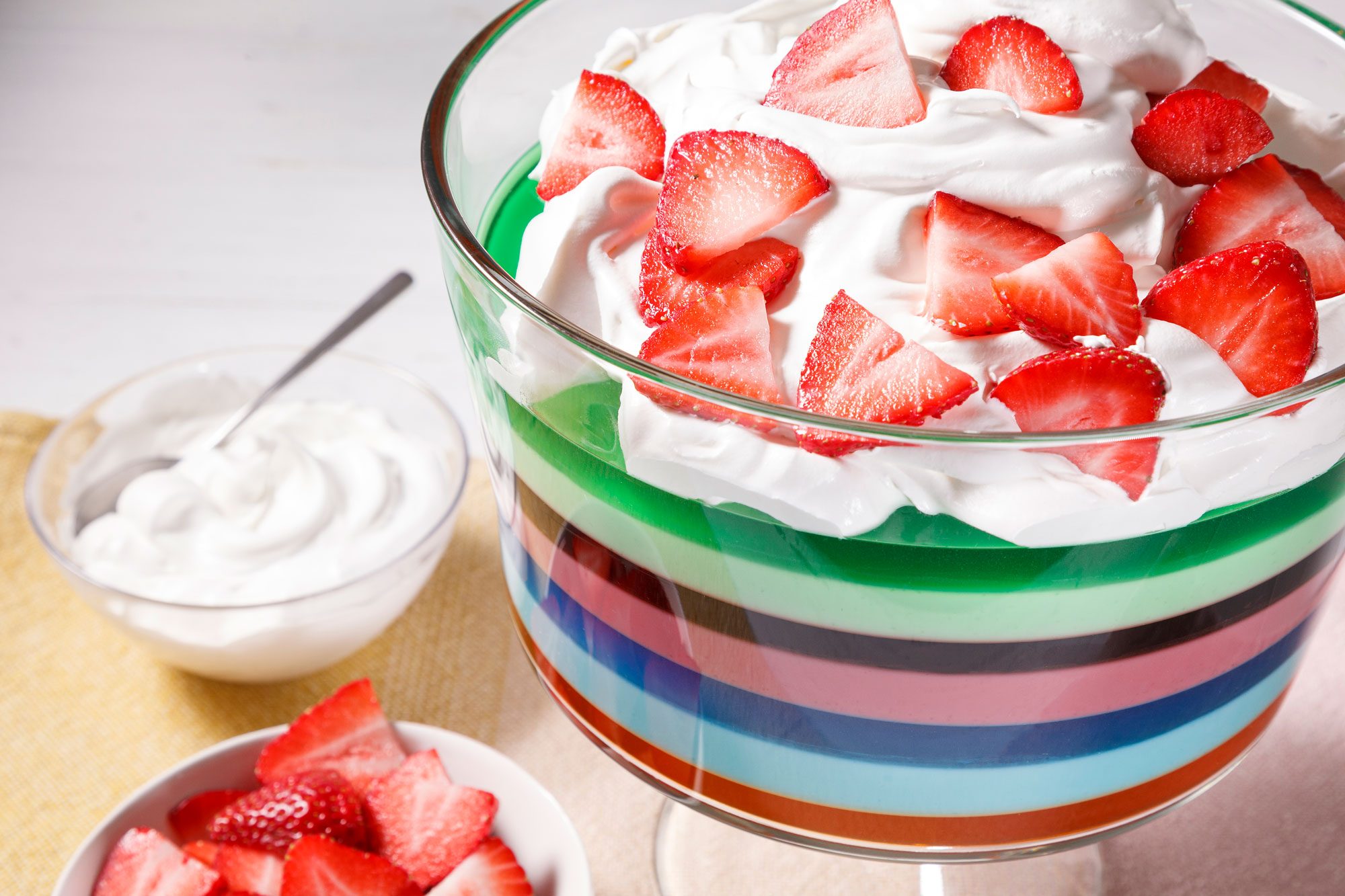 A clear glass bowl filled with a layered dessert consisting of green , pink , blue and orange colored jelly with a layer of whipped cream topped with sliced strawberries