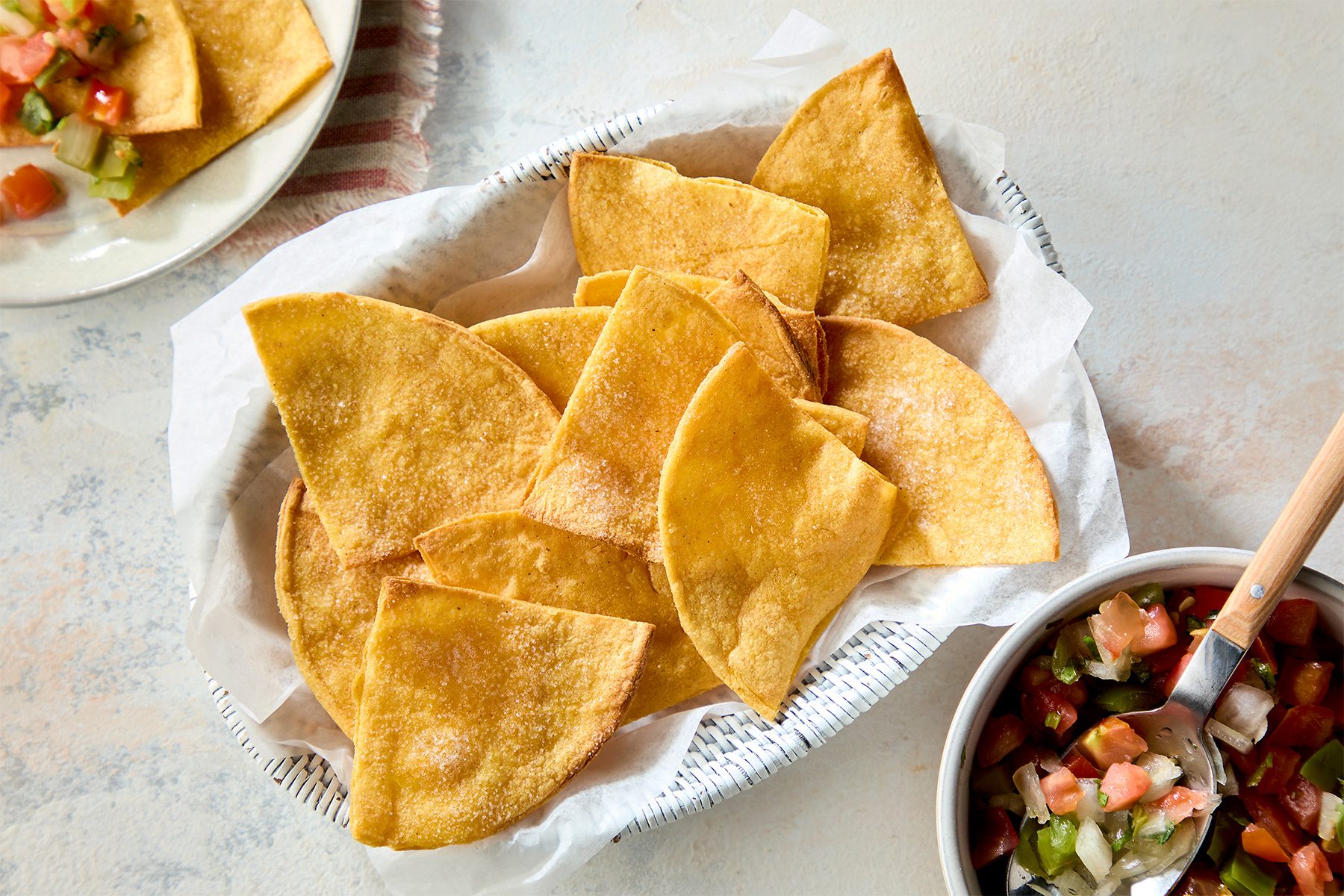 overhead shot of a basket of tortilla chips, a bowl of salsa, and a wooden spoon, The chips are piled high and appear to be lightly salted, The salsa is a vibrant red and green, and the spoon is resting on top of the bowl;