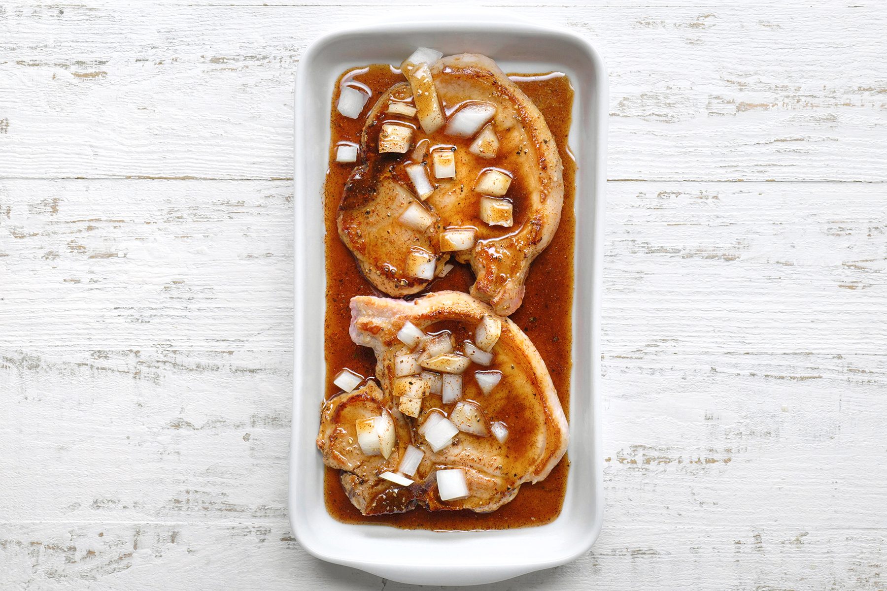 Two seasoned pork chops sit in a white rectangular dish, marinated with a sauce and topped with chopped onions. The dish is placed on a textured white wooden surface.