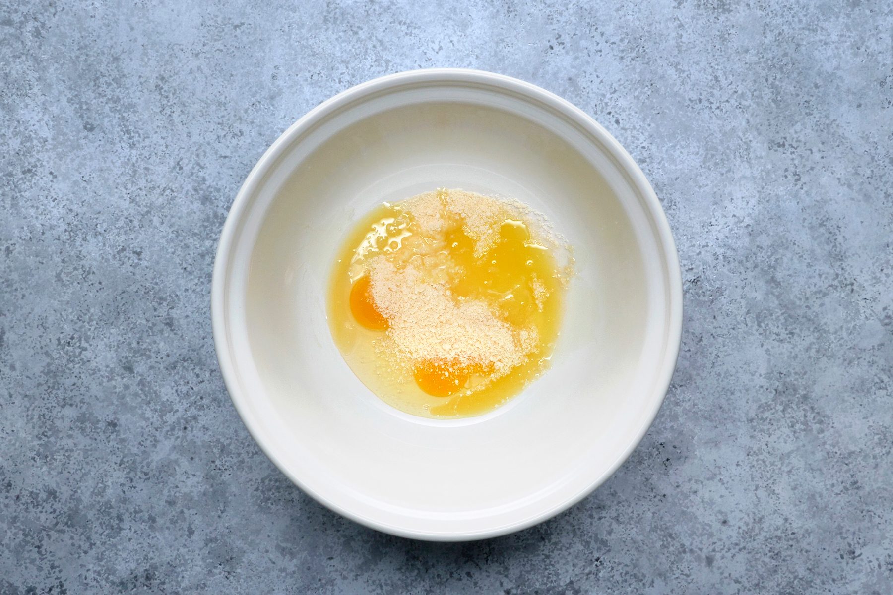 A white bowl with two cracked eggs and some breadcrumbs on a gray, textured surface.