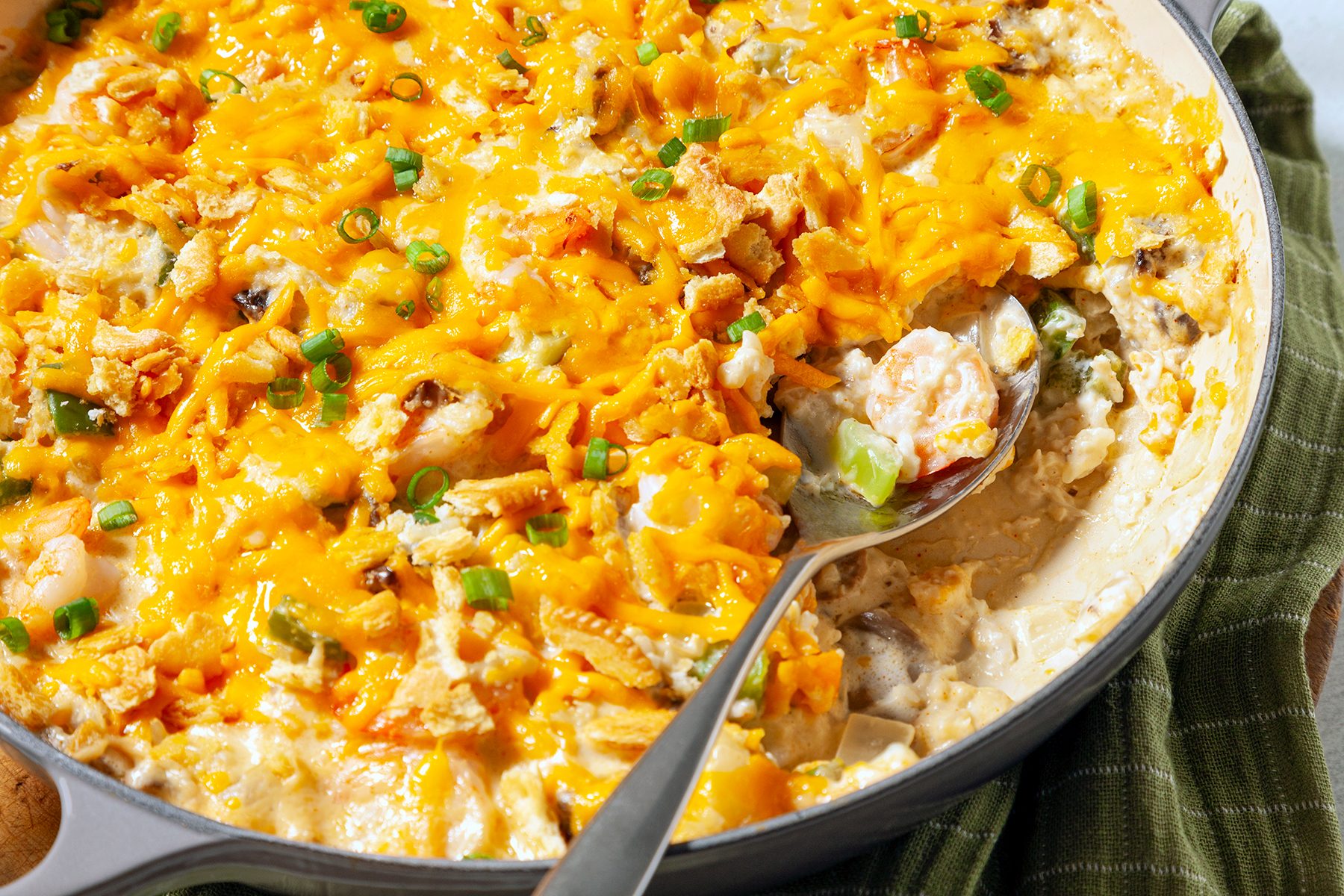 A cheesy casserole with a golden-brown top is garnished with chopped green onions in a skillet. A serving spoon rests inside, revealing a creamy interior filled with various ingredients. It sits on a green cloth.