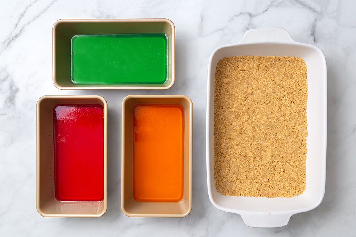 Lime, strawberry and orange gelatin setting in serperate loaf pans and a prepared graham cracker crust in a baking pan for step one of Broken Glass Jello recipe for Taste of Home