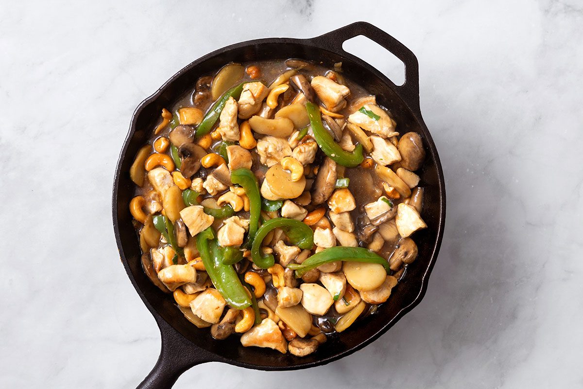 Chopped chicken breasts and salted cashews added to stir fry mixture in cast iron skillet for step three of Cashew Chicken recipe for Taste of Home