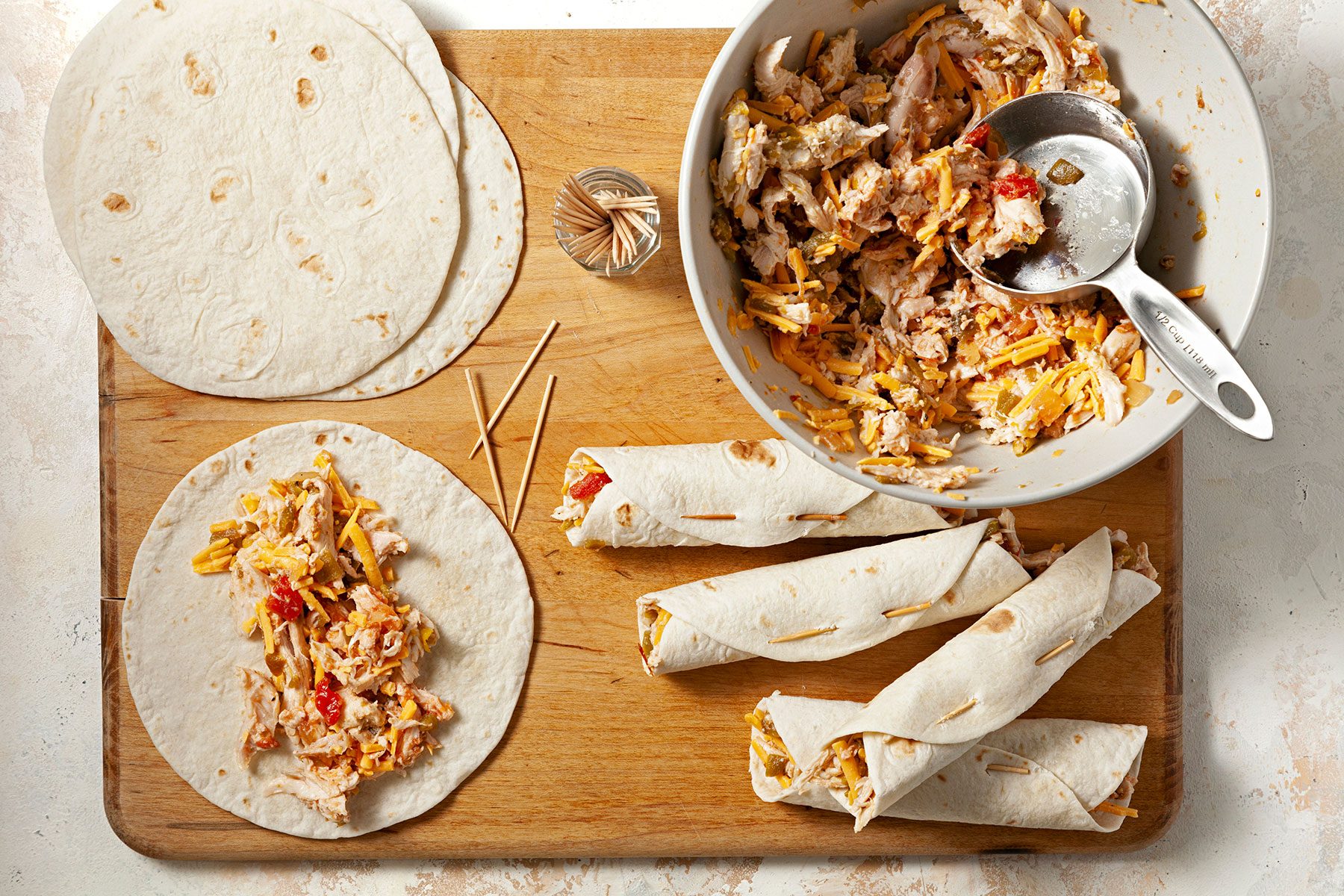A wooden board displays tortillas being filled with a chicken, cheese, and pepper mixture. Some tortillas are rolled and secured with toothpicks, while others and the filling in a bowl. A metal scoop rests in the bowl.