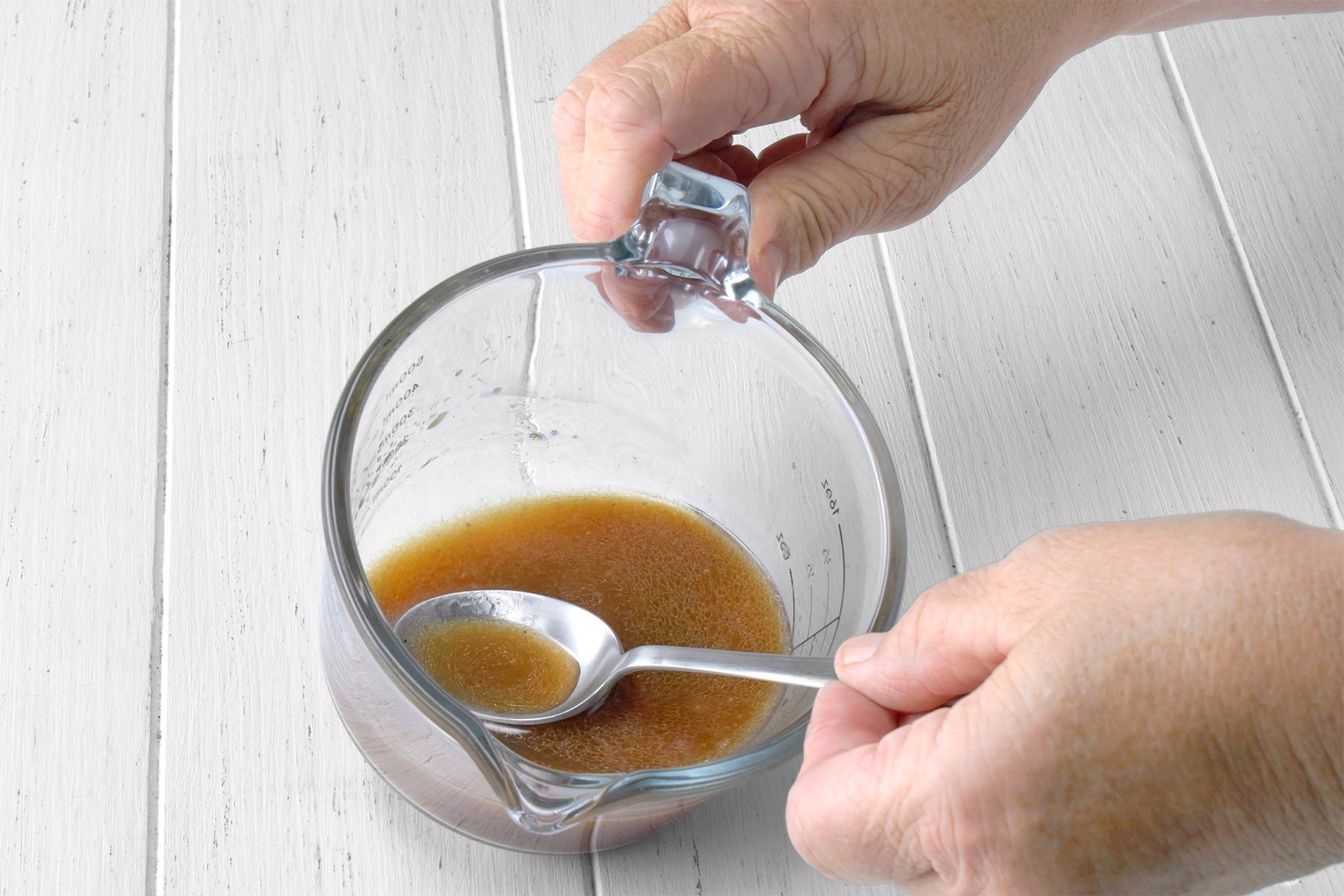 3/4th shot of a person stirring a mixture in a glass measuring cup, The mixture is a dark brown color and has a thick consistency, The person is using a spoon to stir the mixture, and the spoon is partially submerged in the liquid, The measuring cup is sitting on a white wooden surface;