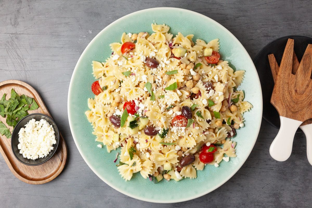 Full yield of Taste of Home Chickpea Pasta Salad in a light blue serving bowl, wooden salad servers, feta and basil