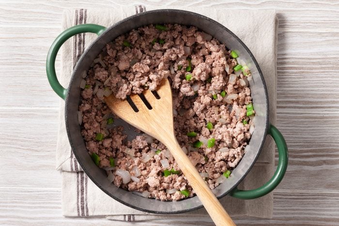 An overhead shot of a green cast-iron pot on a wooden surface. The pot contains cooked and crumbled ground meat, likely ground beef, mixed with chopped green onions. A wooden spatula rests in the pot, and a kitchen towel is placed underneath.