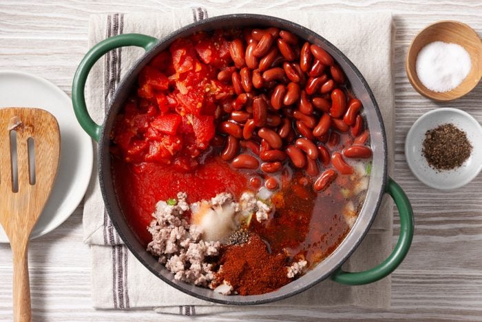 An overhead shot of a green cast-iron pot filled with various ingredients for a chili recipe. Ground meat, diced tomatoes, kidney beans, and a liquid base are visible in the pot. A wooden spatula rests on the rim, and a small bowl of salt and a bowl of ground black pepper are placed nearby.