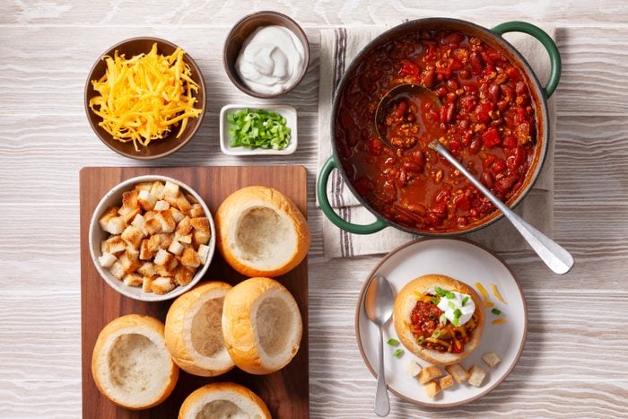 An overhead shot of a delicious and hearty chili dish, perfect for a cozy meal. A large green Dutch oven filled with thick, red chili serves as the centerpiece of the image. The chili is loaded with ground beef, beans, and vegetables, garnished with shredded cheese and chopped green onions. Surrounding the chili are several hollowed-out bread bowls, ready to be filled with the flavorful chili. Additional ingredients, such as shredded cheese, sour cream, and croutons, are also present.