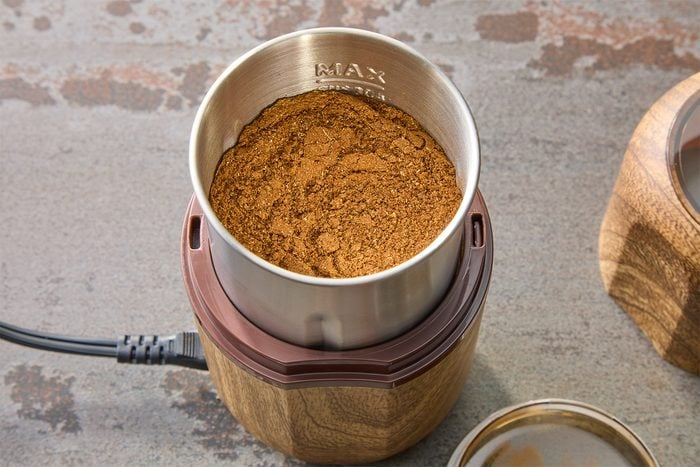 A metal cup filled with ground coffee sits inside a wooden base of a coffee grinder. The grinder has a power cord attached. The surface underneath is textured and gray.