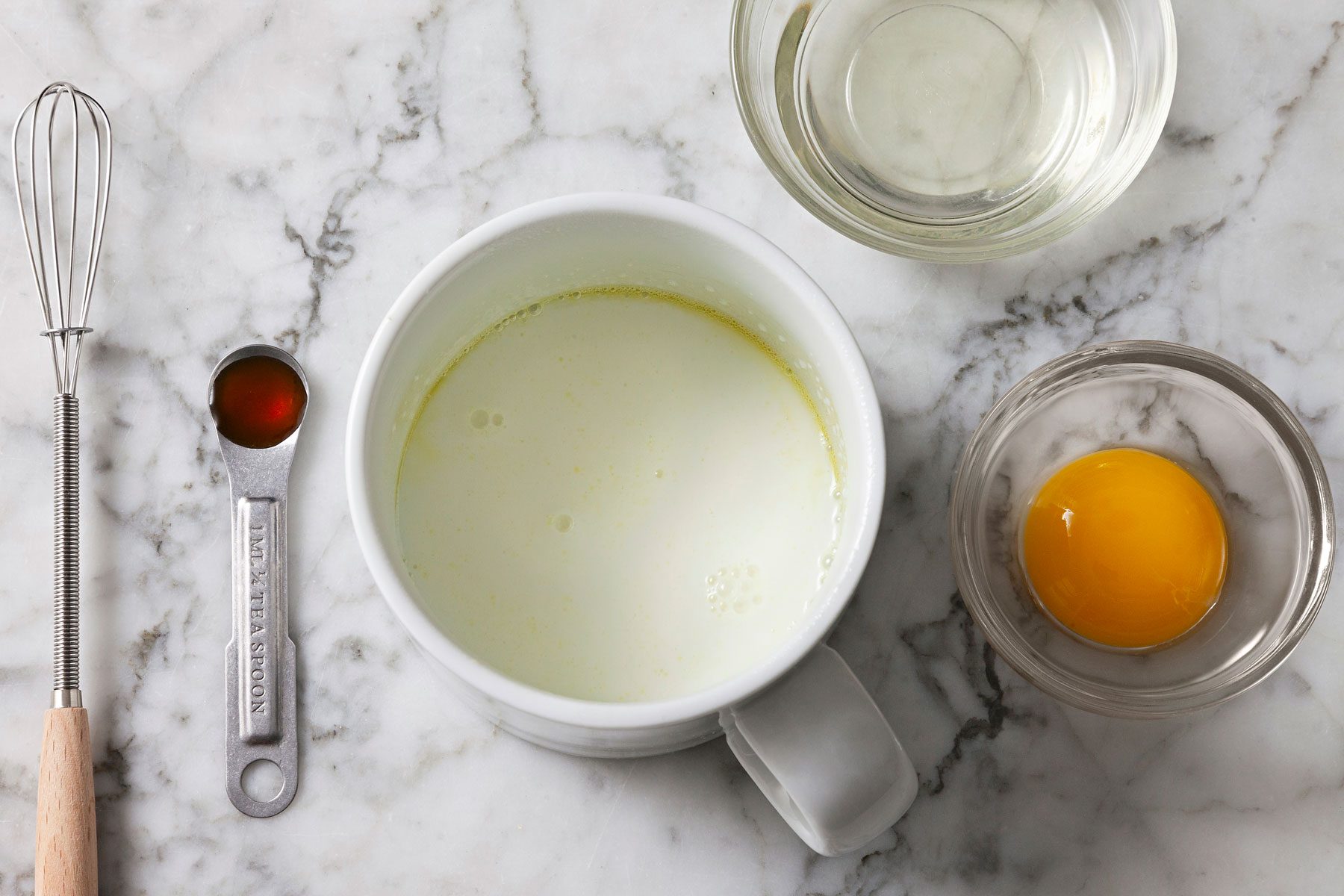 Milk and oil combined in a mug with egg yolk and vanilla extract next to it.