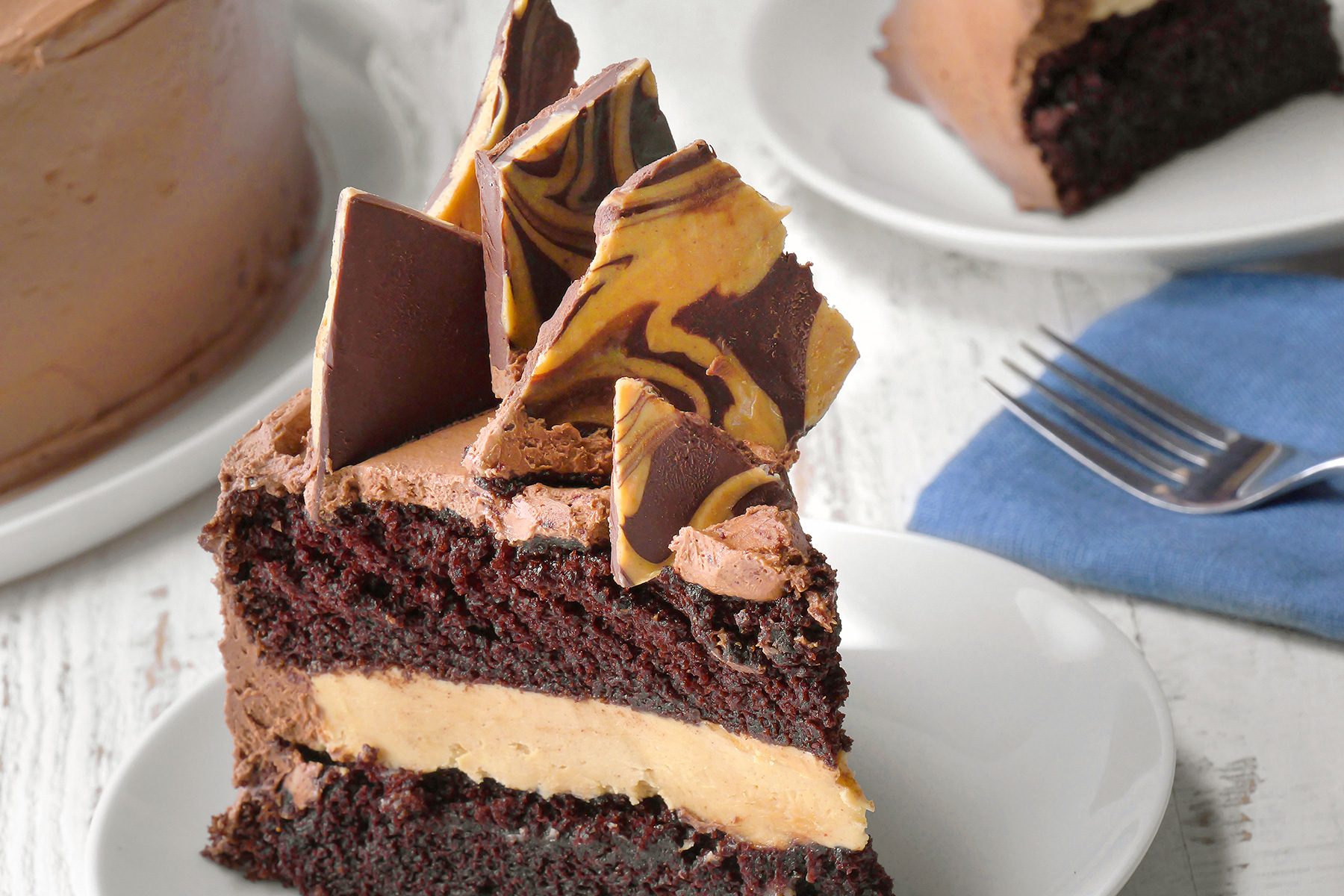 A slice of decadent chocolate cake with layers of creamy filling and topped with decorative pieces of marbled chocolate sits on a white plate. Nearby, a fork rests on a blue napkin, and part of another cake is visible in the background.