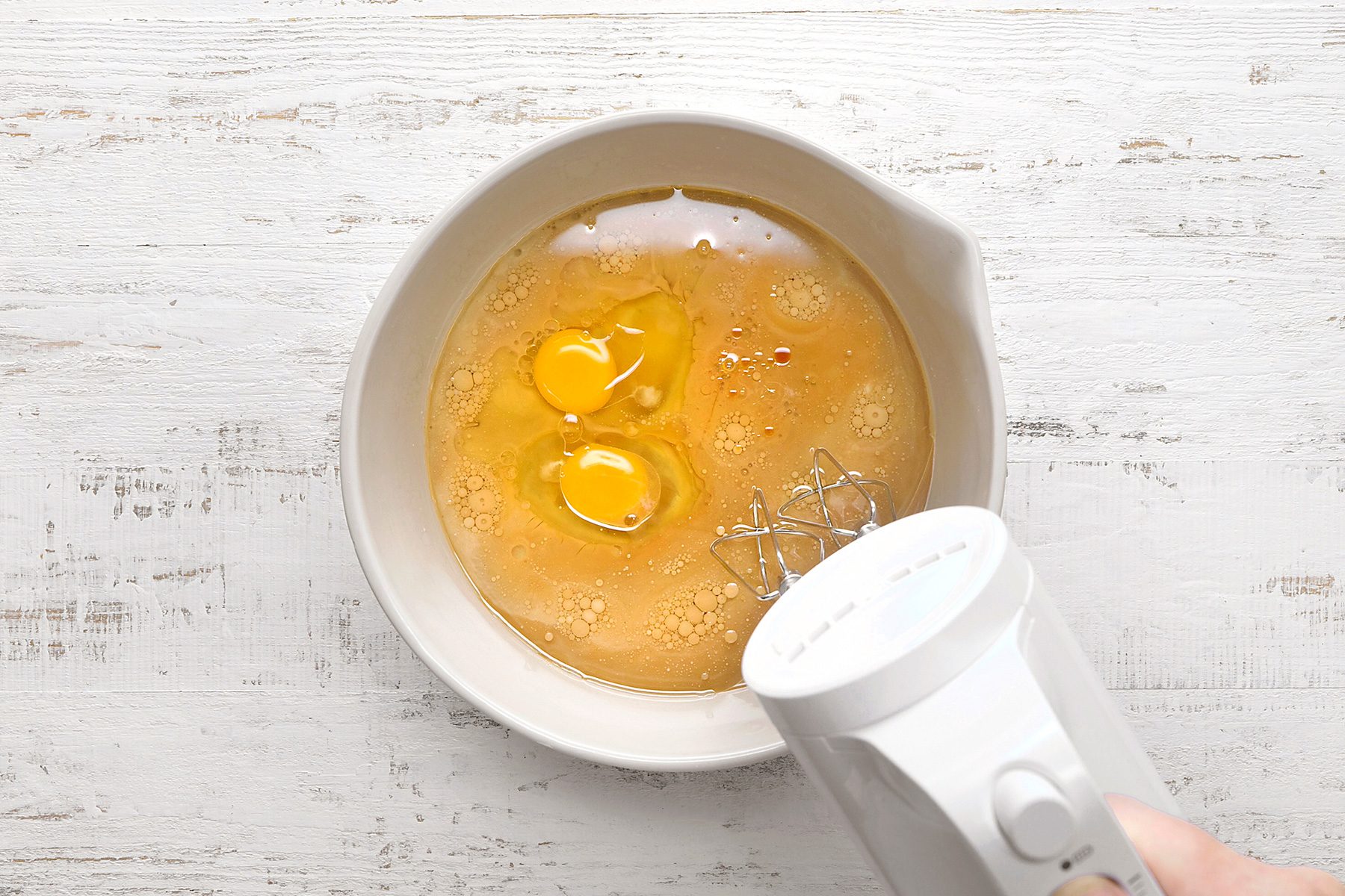 A top-down view of a white mixing bowl containing two cracked eggs and other ingredients. A white electric hand mixer is partially visible on the right, hovering over the bowl, ready to mix the contents. The background is a white wooden surface.