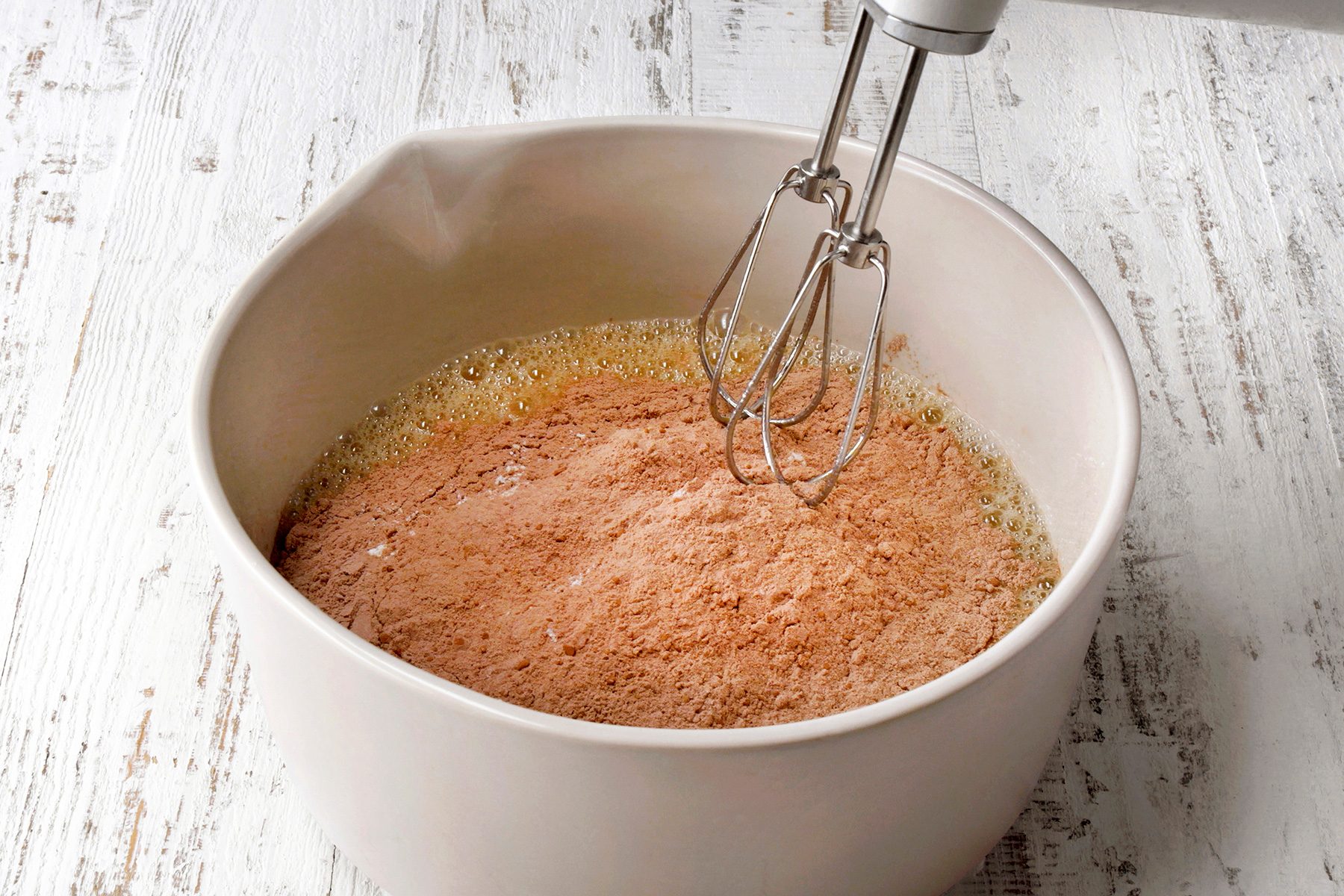 A white mixing bowl on a light wooden surface contains a mixture of dry chocolate cake mix and wet ingredients. An electric mixer with twin beaters is placed in the bowl, ready to blend the ingredients.
