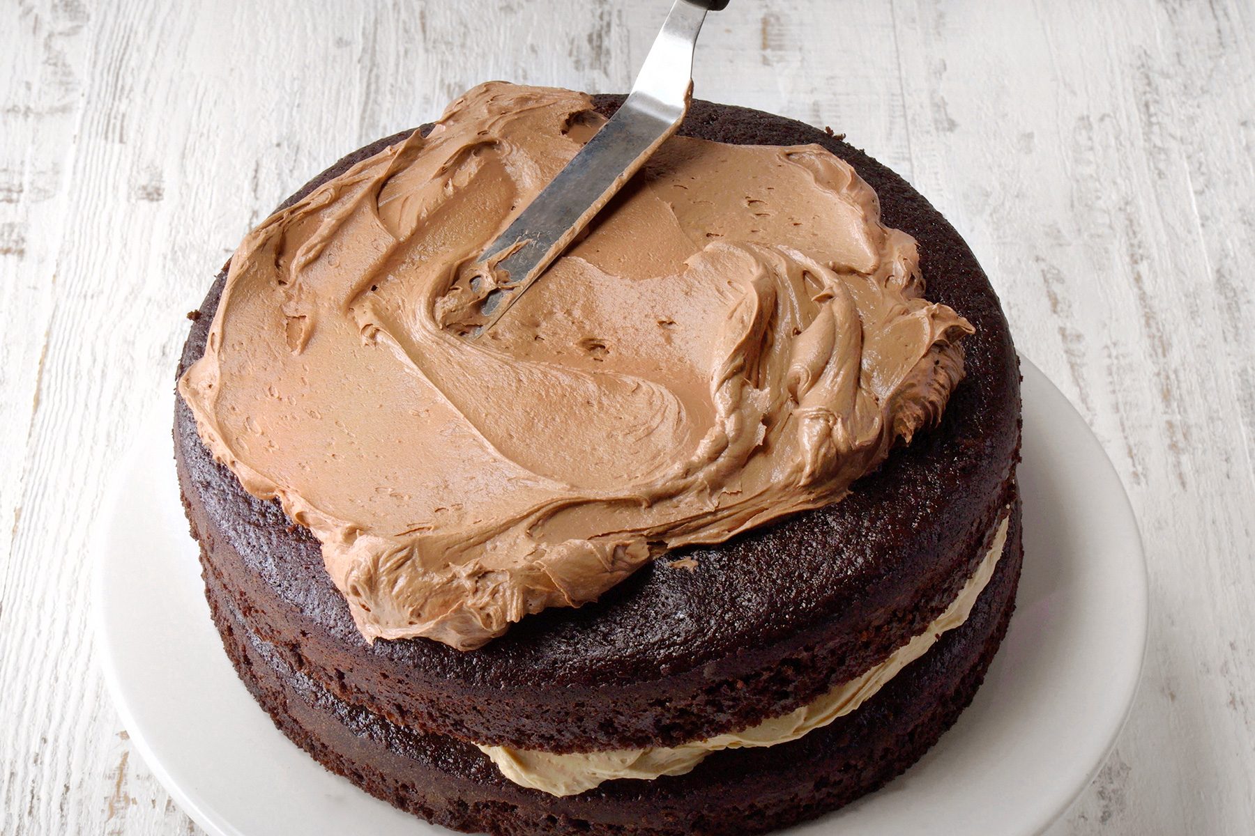 A layered chocolate cake being frosted with light brown icing on a white plate. The cake sits on a rustic white wooden surface. A metal spatula is smoothing the frosting on the top layer.