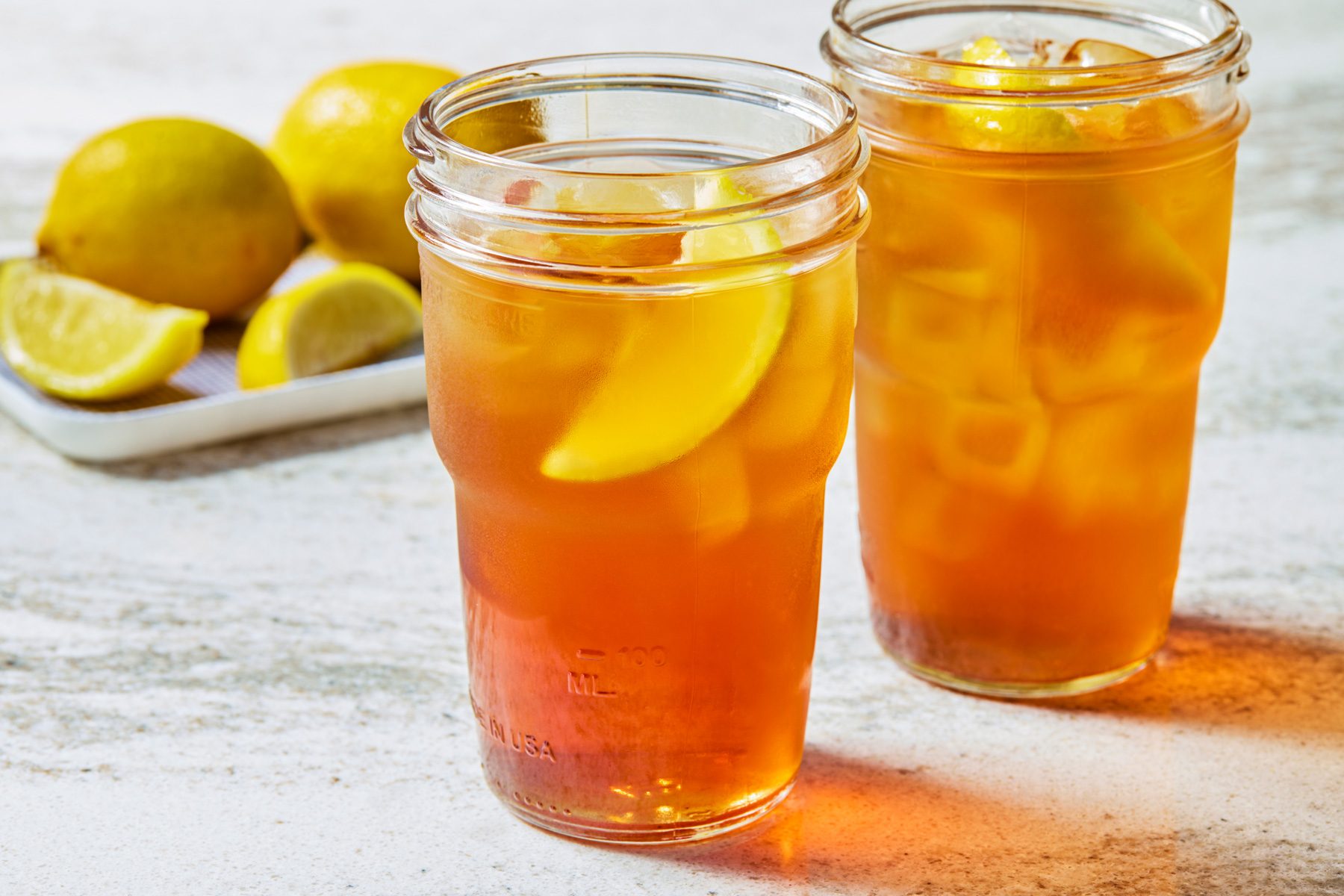 3/4 shot of two mason jars filled with iced tea and lemon wedges, placed on a white surface, with additional lemon wedges and a white plate in the background.