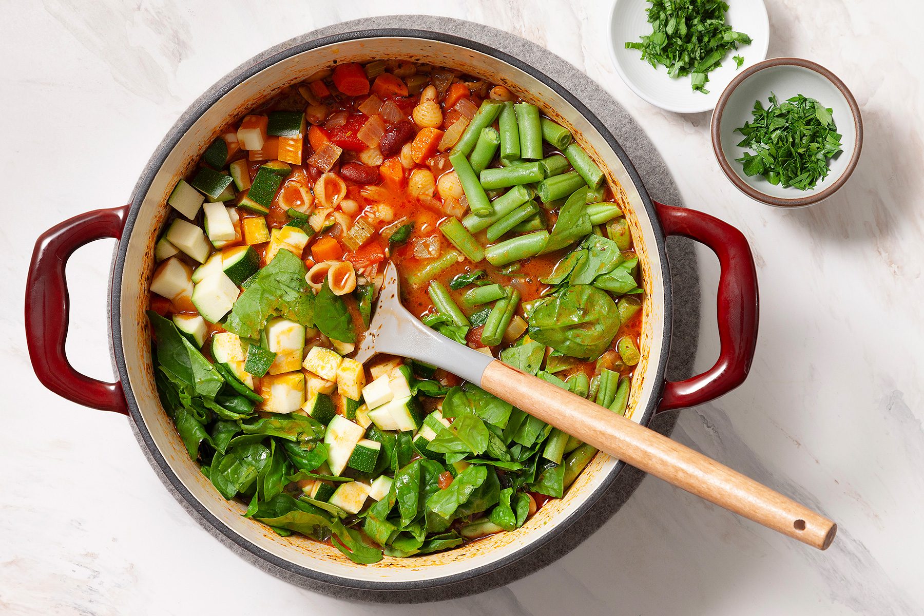 A pot of vegetable stew filled with green beans, spinach, zucchini, and a tomato-based broth. A wooden spoon rests inside the pot. Chopped herbs are in two small bowls nearby on the white countertop.