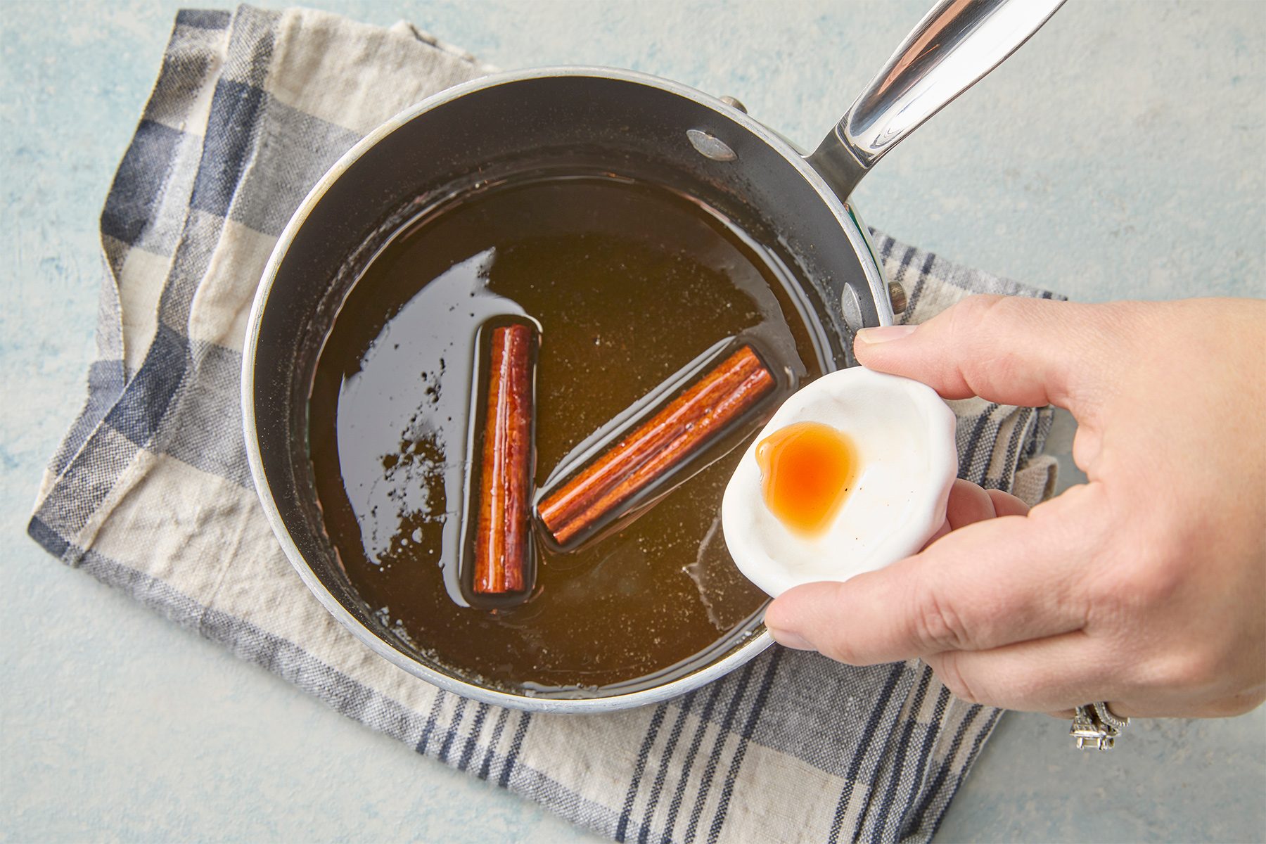 A hand holding a small bowl of vanilla extract above a saucepan with syrup and cinnamon sticks on a checkered cloth.