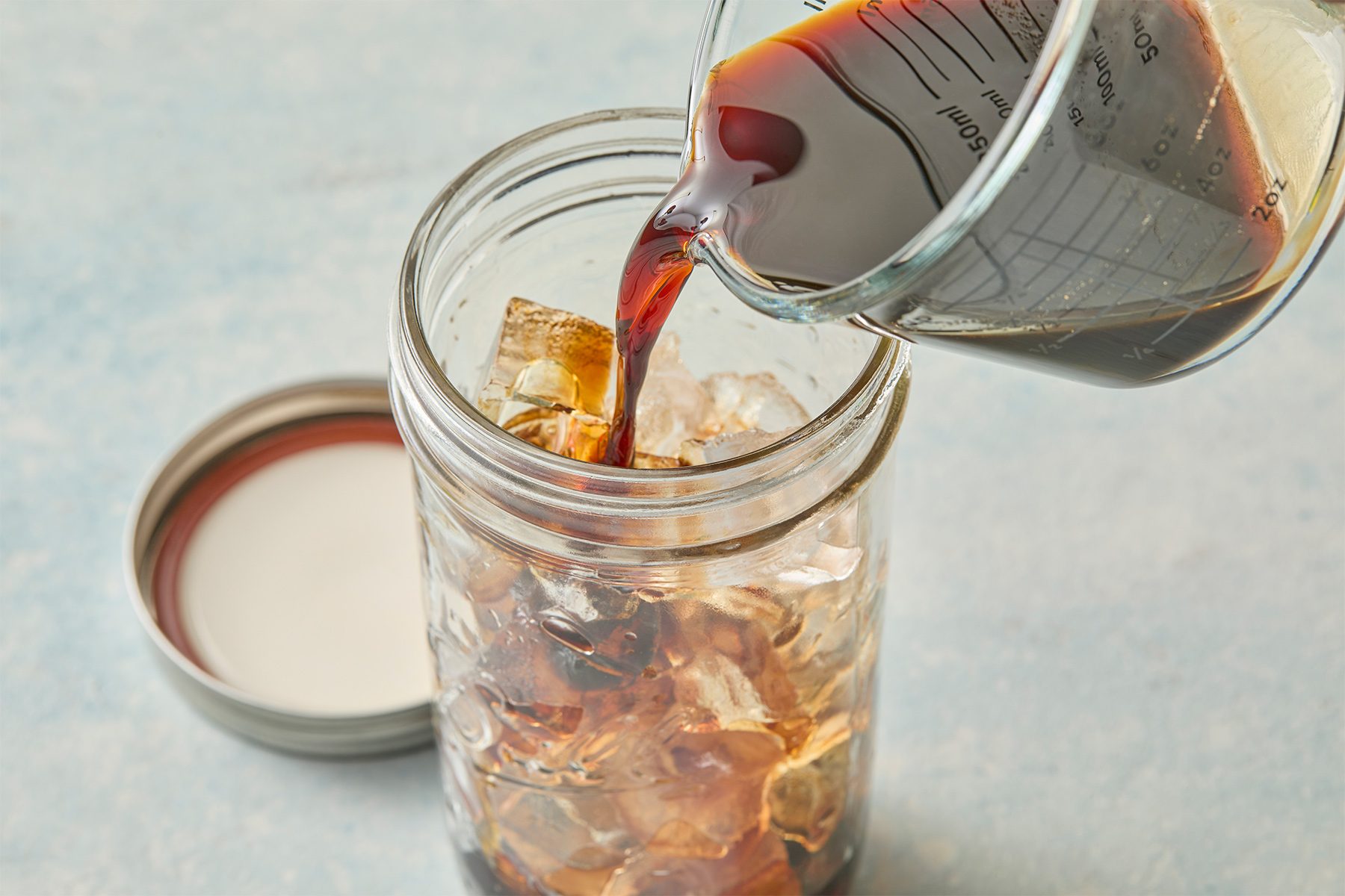 A close-up of cold brew coffee being poured from a measuring cup into a glass jar filled with ice. The lid of the jar is on the table to the side, and the background is a light, textured surface.