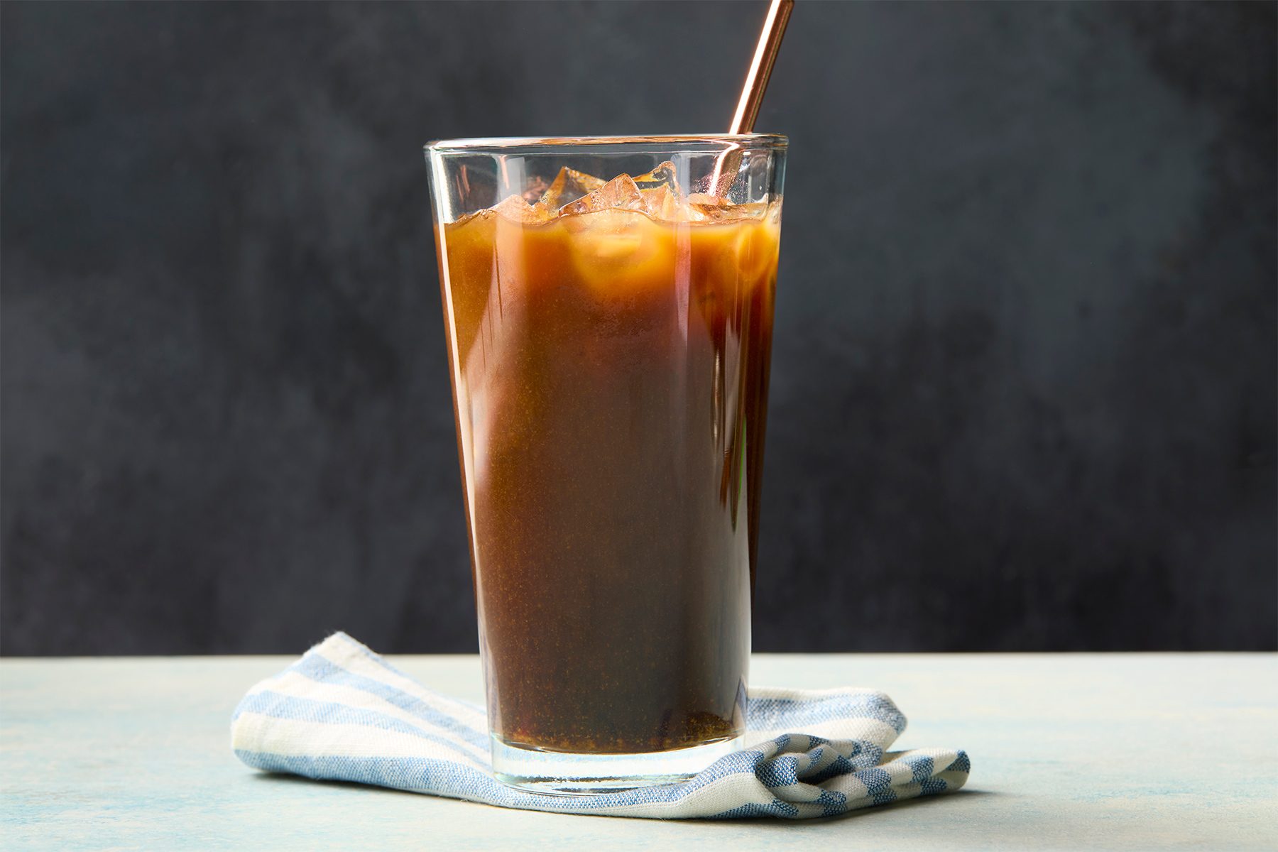 A glass of iced coffee with a metal straw sits on a folded, blue and white checkered cloth. The background is a textured, dark surface.