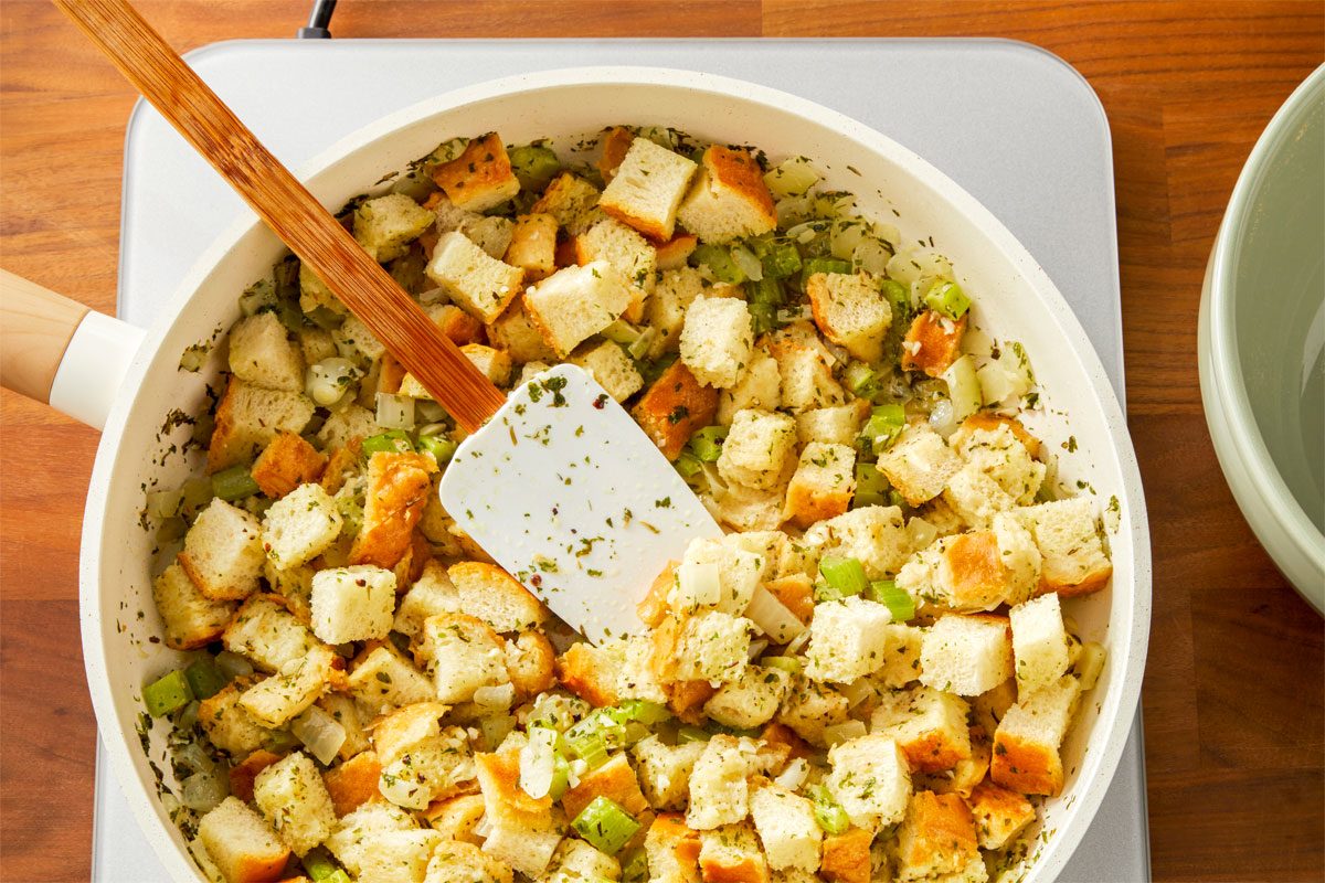 overhead shot of a white pan filled with a mixture of diced bread, chopped celery, and chopped onions, The bread cubes are a light brown color, and the vegetables are a mix of green and white, A wooden spatula is being used to stir the ingredients together