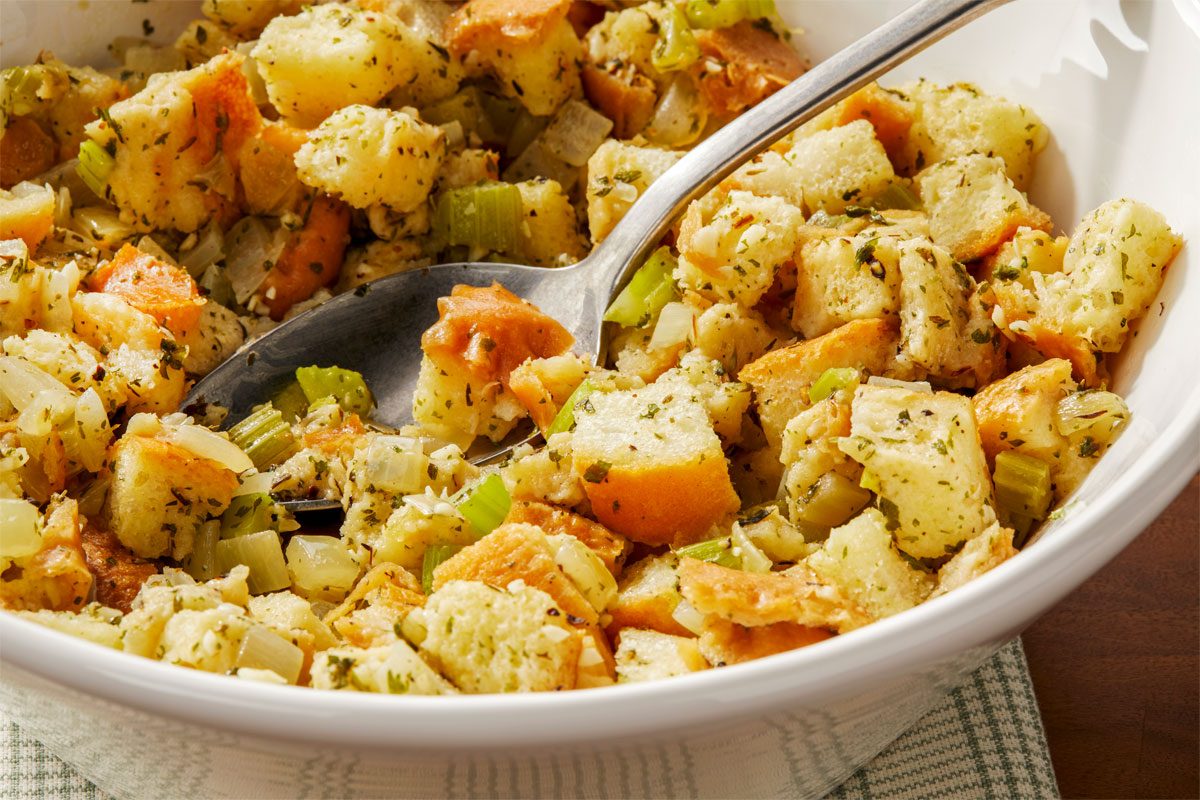 shot of a white bowl filled with a mixture of diced bread, chopped celery, and chopped onions, The bread cubes are a light brown color, and the vegetables are a mix of green and white, The mixture is seasoned with herbs and spices, bowl is placed over white kitchen cloth, A spoon is resting in the bowl;