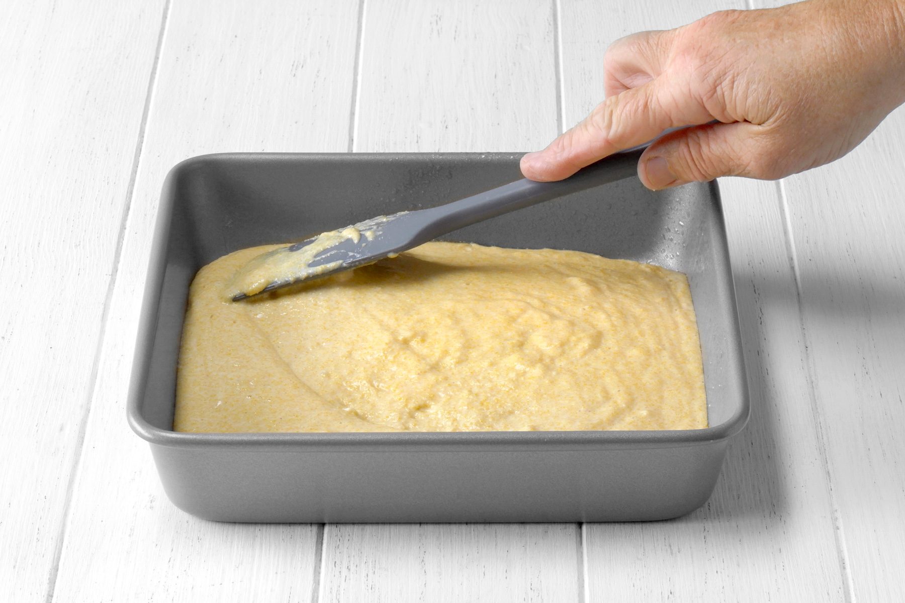 3/4th shot of a hand using a spatula to spread a batter evenly in a square baking pan, The batter is a light yellow color and has a smooth texture, The baking pan is gray and is sitting on a white wooden surface;