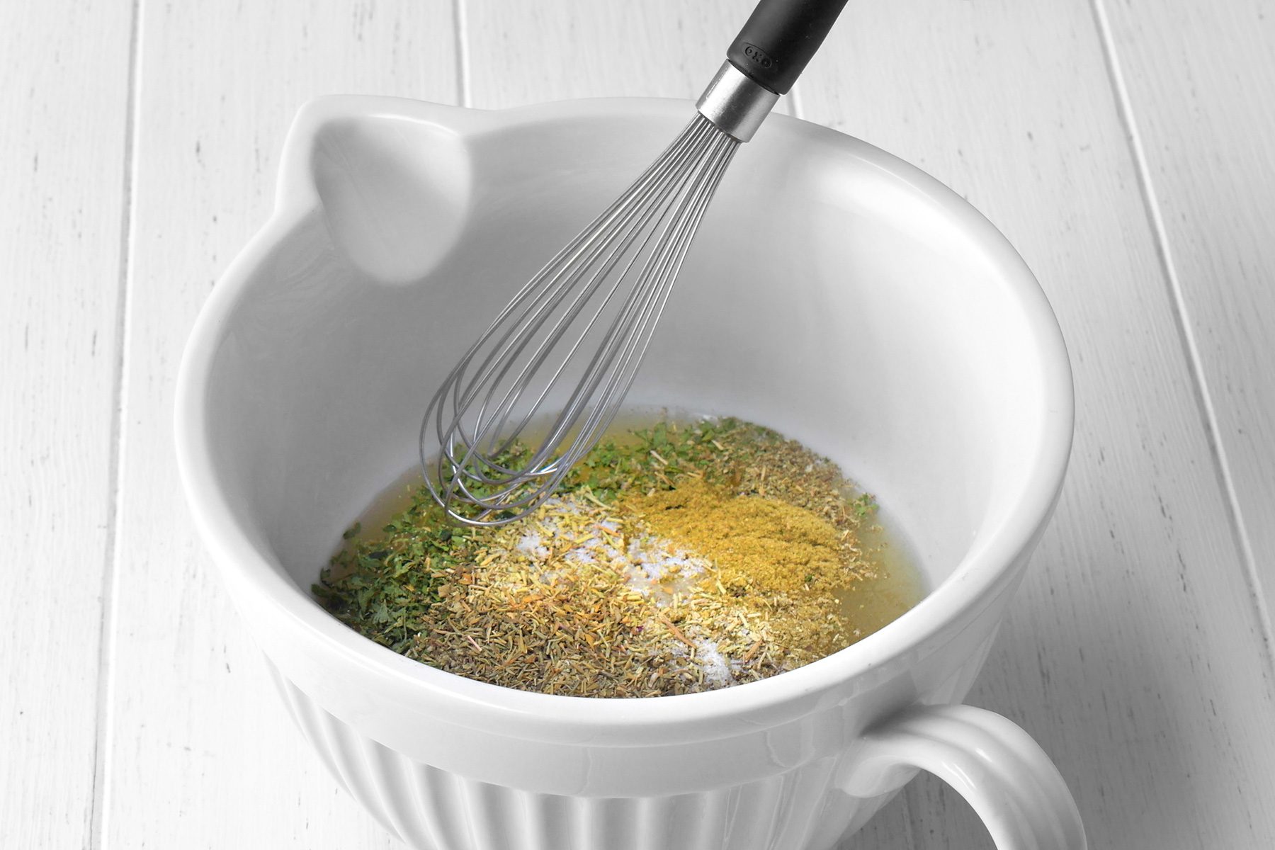 3/4th shot of a white bowl contains a mixture of liquid and various spices, The liquid appears to be a light yellow color, and the spices are a variety of colors, including brown, white, and green, A whisk is being used to stir the mixture together, The bowl is sitting on a white wooden surface;