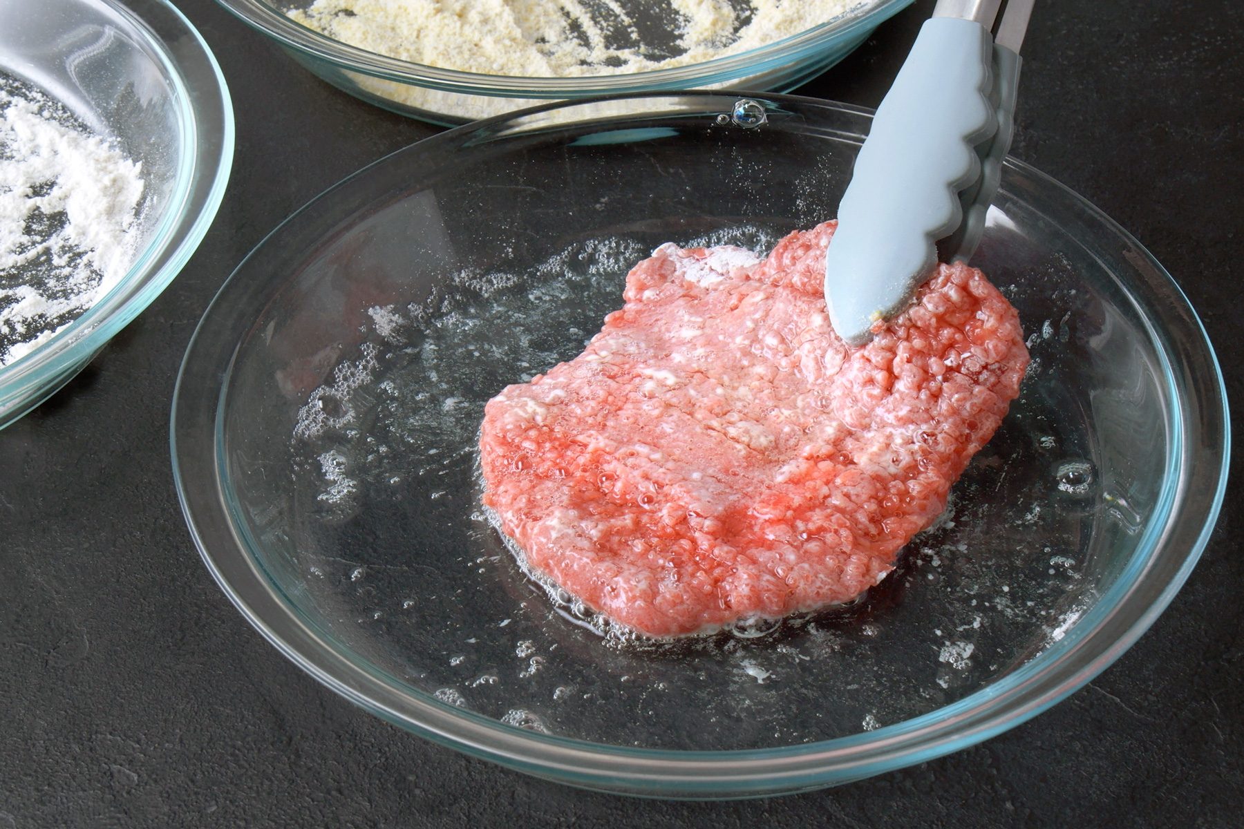 3/4th shot of a breaded meat patty being dipped into a dish of whisked eggs, The meat patty is coated in flour and is being submerged in the egg mixture, The egg mixture is a light yellow color and has bubbles forming on the surface