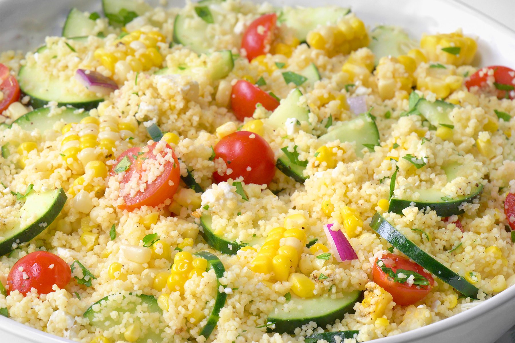 close shot of a white bowl is filled with a colorful couscous salad, The salad contains couscous, diced cucumbers, cherry tomatoes, crumbled feta cheese, and chopped fresh parsley, The ingredients are mixed together with a light dressing;
